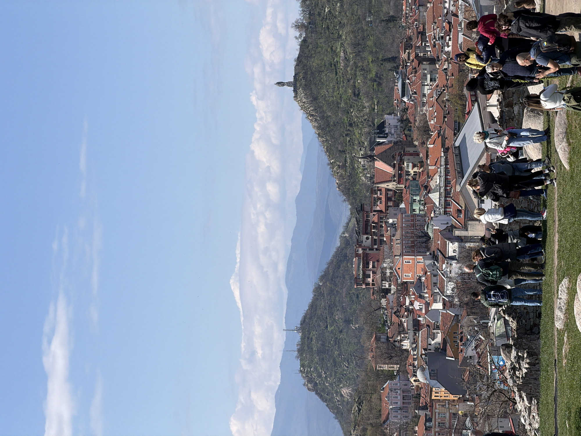 The image shows a panoramic view from Nebet Tepe in Plovdiv, Bulgaria. The sky is blue with scattered clouds on the left. In the center are tree-covered hills and distant mountains. On the right is a dense urban landscape with buildings featuring red tile roofs. A group of people stand on the edge of Nebet Tepe.
