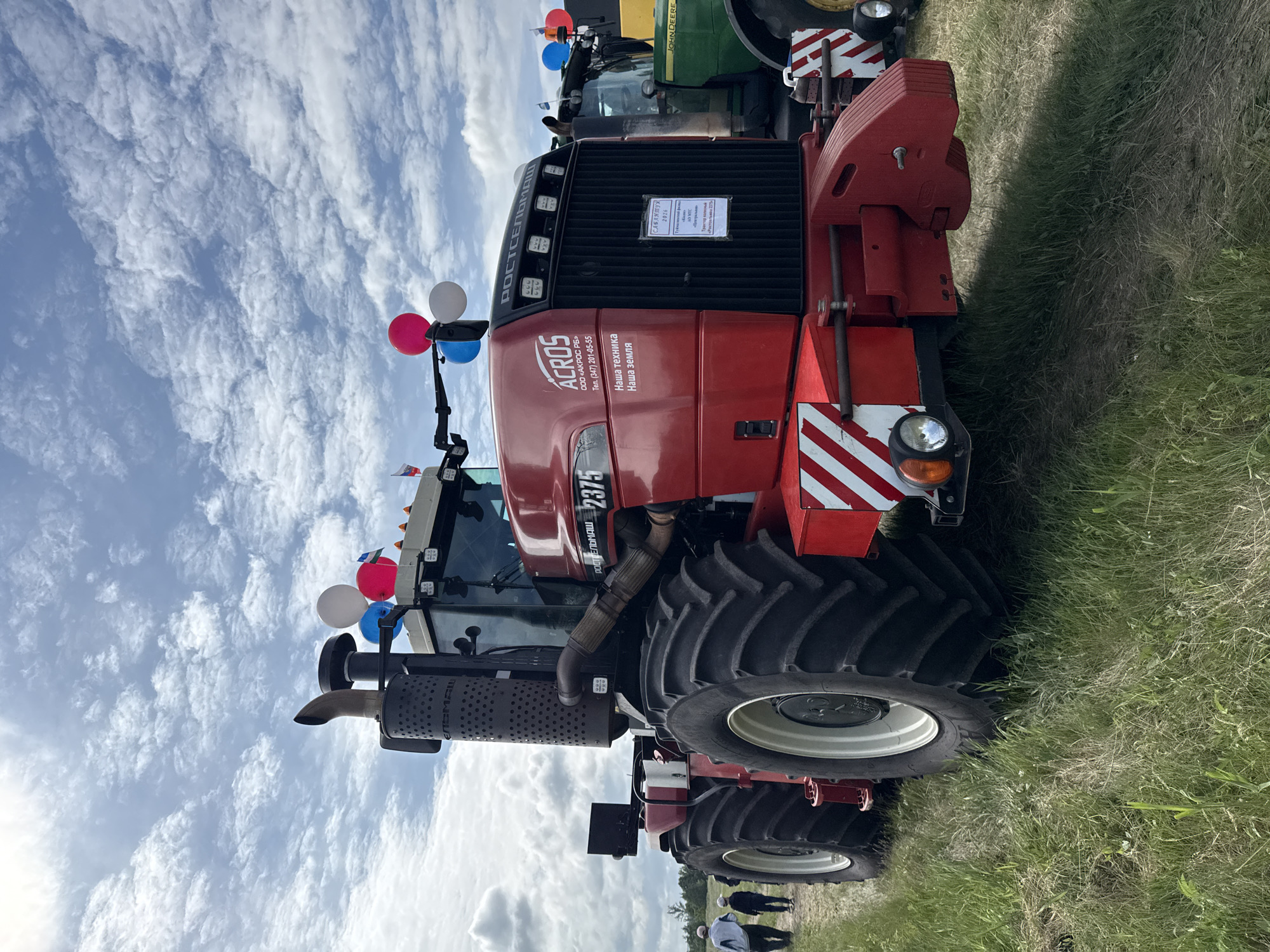 The image shows a red tractor with the model number 2375 and the brand "ACROS". It is parked on a grassy field under a cloudy sky. Several balloons are attached to the top of the tractor. In the background, another tractor, a green John Deere, is visible. Two people, likely attendees, are walking in the distance towards the left side of the frame. The tractor appears to be part of an agricultural exhibition or event. The location of Norkeevo, Russia, is not visually evident in the image itself.