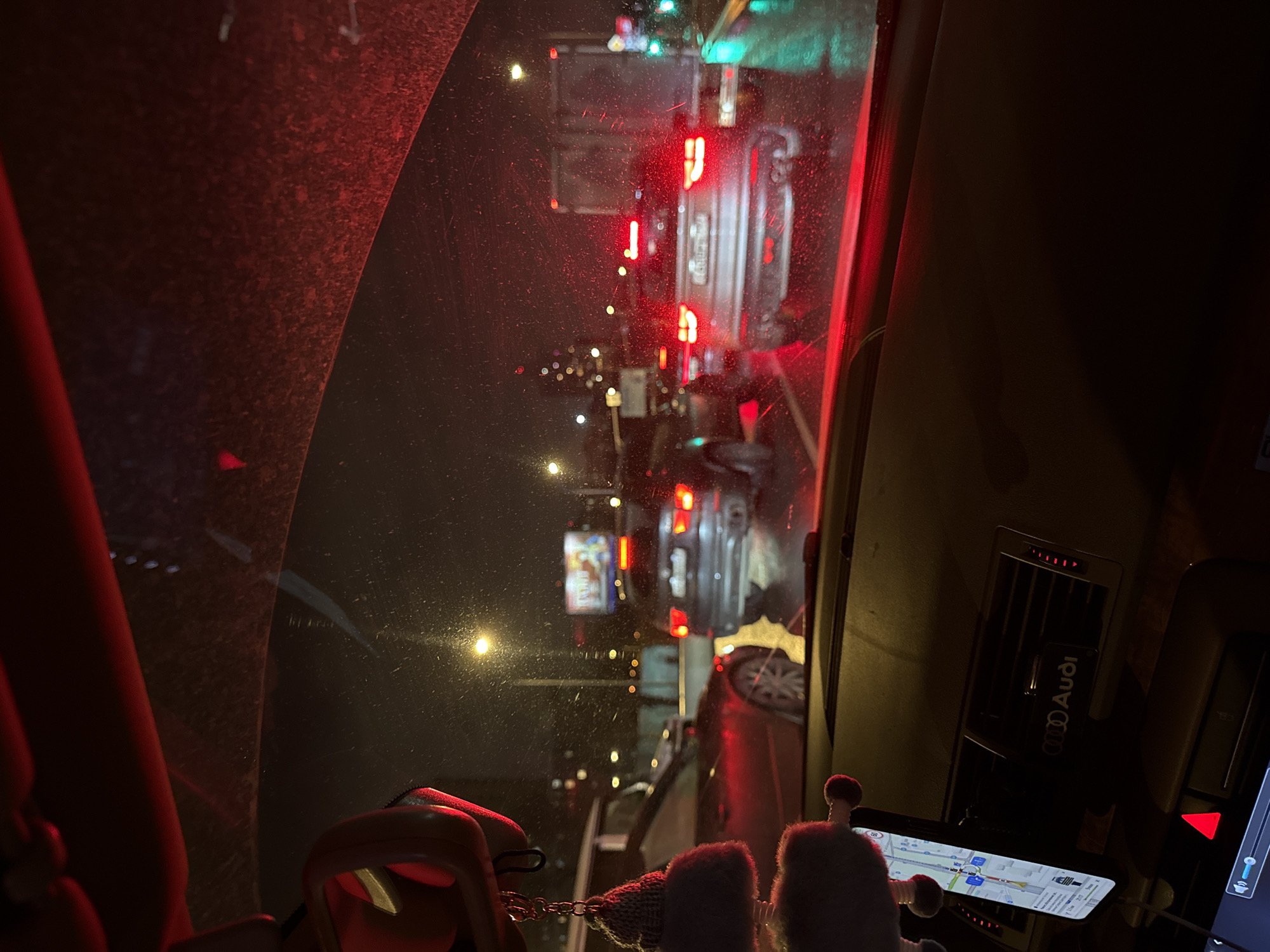 The image, taken from inside a car, shows a traffic jam in Moscow, Russia. The windshield has streaks, possibly from rain or cleaning. A line of cars with lit red taillights stretches into the distance, illuminated by streetlights and the glow of city lights. Inside the car, an Audi logo is visible on a vent, suggesting the make of the vehicle. A navigation app is open on a smartphone mounted on the dashboard. There are two fuzzy figures on the dashboard.