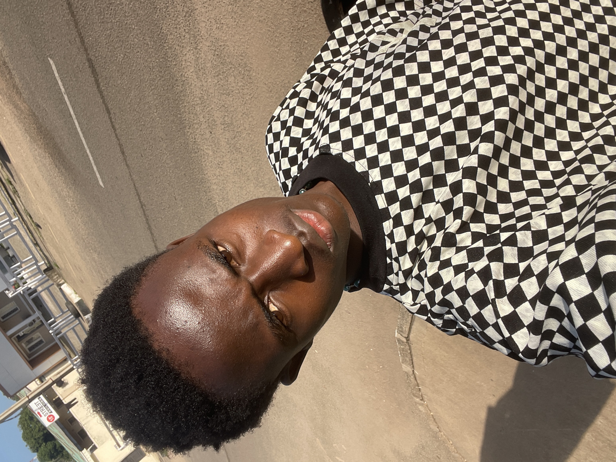 In an outdoor, sunlit environment on a street in Jos, Nigeria, a young Black male is captured in a close-up, high-angle selfie. He is positioned at the center of the frame, with his head tilted to his left and his gaze directed towards the camera. His skin is dark and glistening with sweat, reflecting the bright sunlight. His hair is short and black, styled in a textured afro. He wears a black and white checkered long-sleeved shirt with a black collar. In the background, a paved road with a white line marking the lane is visible, along with the side of a building, a white fence, and some trees. The lighting suggests it is daytime, possibly late afternoon, due to the long shadow cast on the ground to the left of the subject. A sign with red lettering indicating "15 STREET BUILDING" is partially visible on the building in the background.