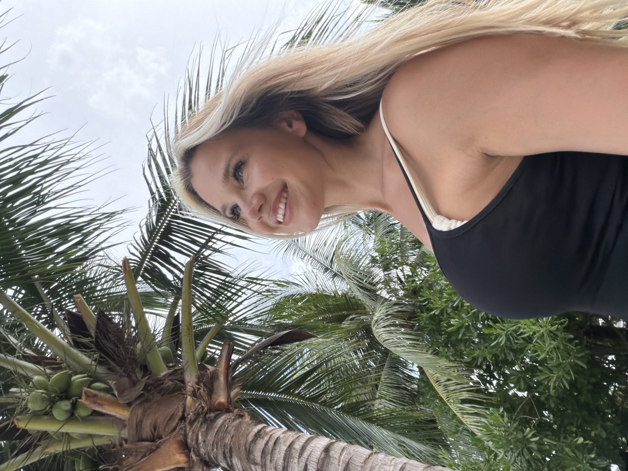 An adult Caucasian female with blonde hair, wearing a black tank top with white trim, stands outdoors in front of palm trees and a cloudy sky. She is looking upwards and to the right with a smile. The image appears to be taken from a low angle. The location is implied to be Wichit, Thailand, consistent with a tropical environment featuring palm trees.