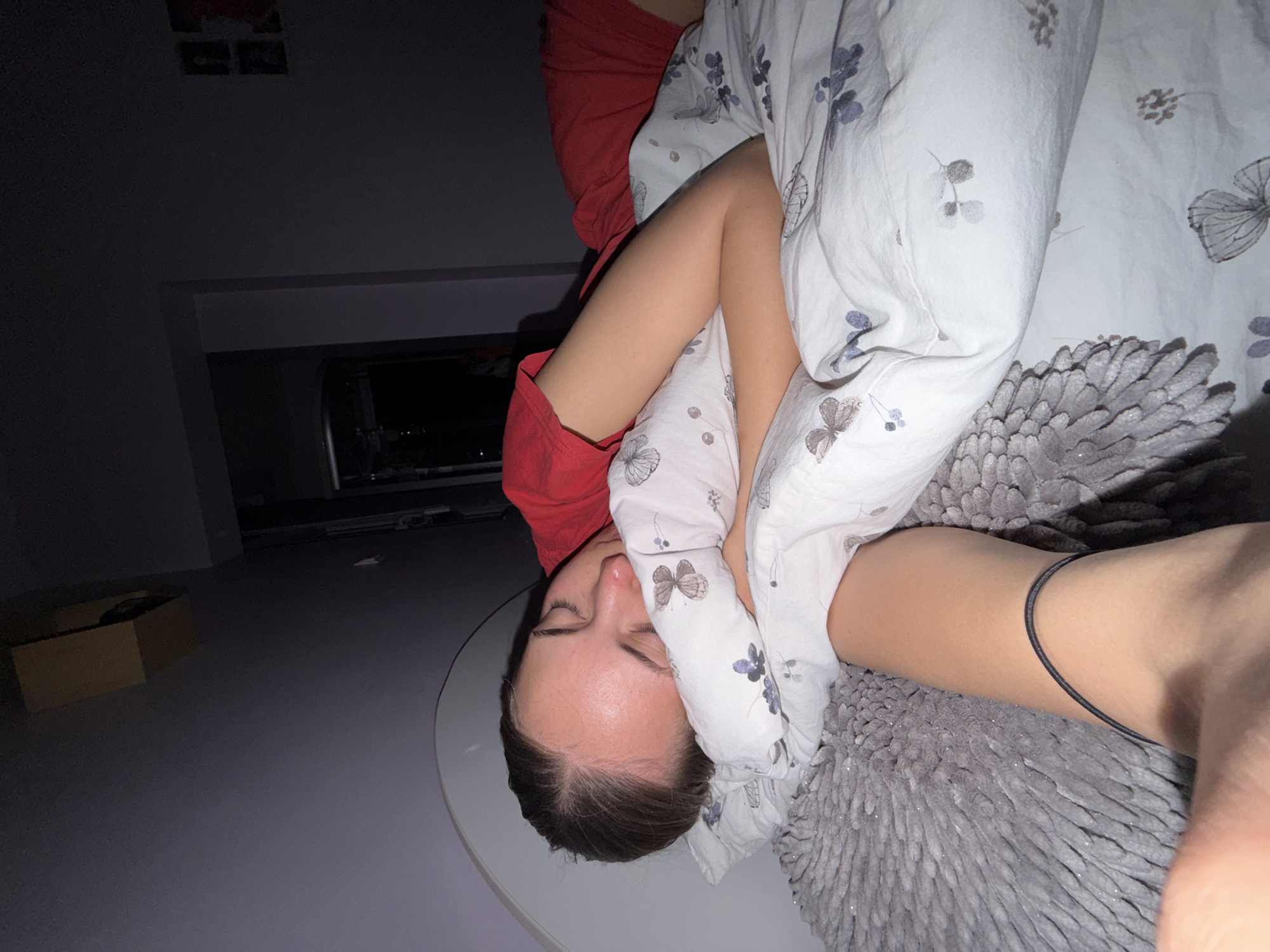 A person lies prone on a white circular surface. The person is partially covered by a white comforter with a floral and butterfly pattern. The person is wearing a red shirt. A textured grey throw is partially visible beneath the comforter. The room is dimly lit, with a cardboard box in the background. A partial view of a garage door is visible in the background, indicating an interior setting. The photograph may be taken in Buhovo, Bulgaria, but no elements of the image definitively verify location.