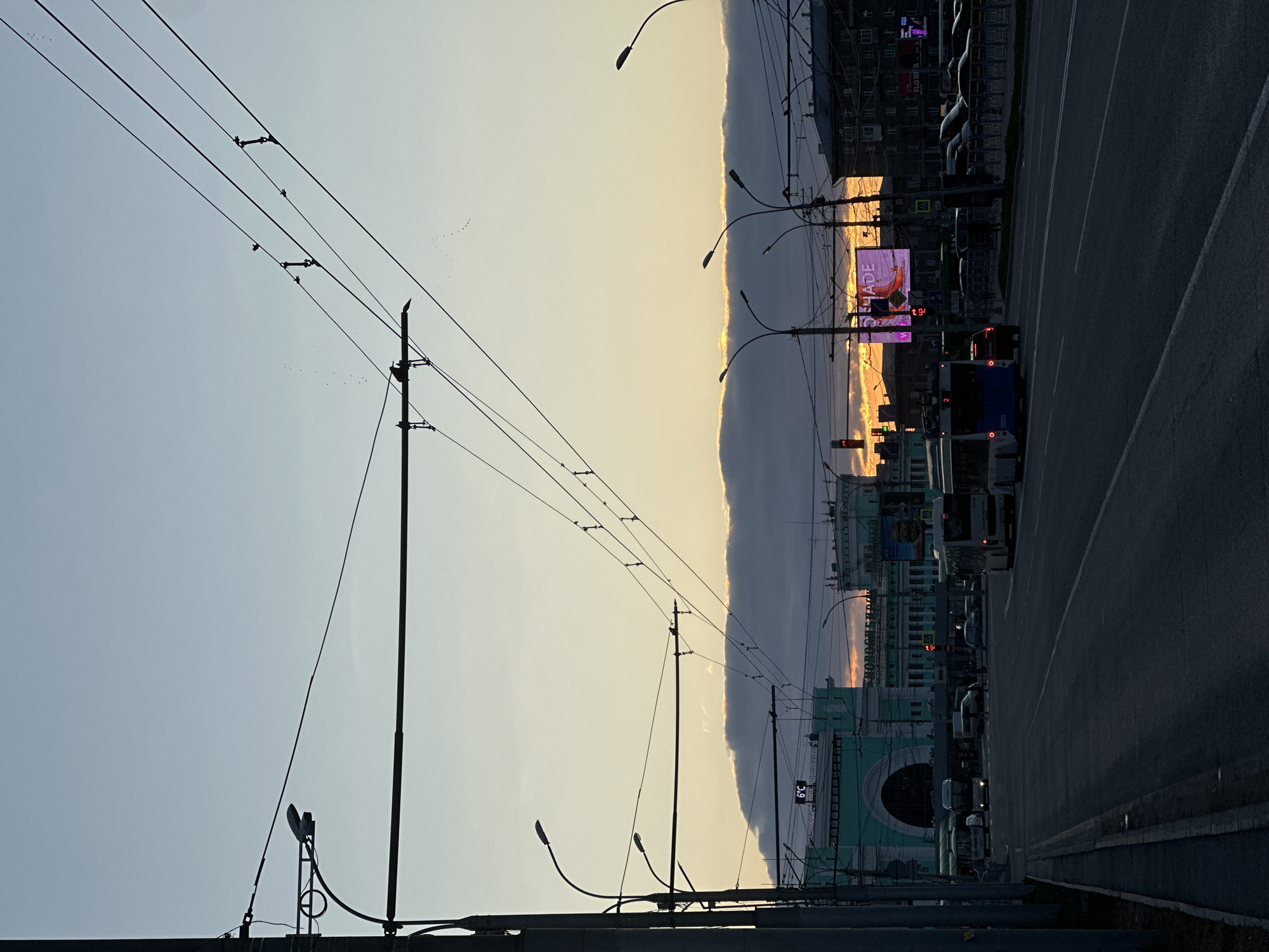 The image shows a street view in Novosibirsk, Russia, taken at dusk. A row of buses are moving on a highway road. A distinctive greenish building with a large arch is visible. Utility poles and power lines run across the sky. A digital billboard advertises "Renade" and another word, possibly referring to a product or brand. The sky has a gradient from light blue to yellowish and orange near the horizon, suggesting a setting sun.