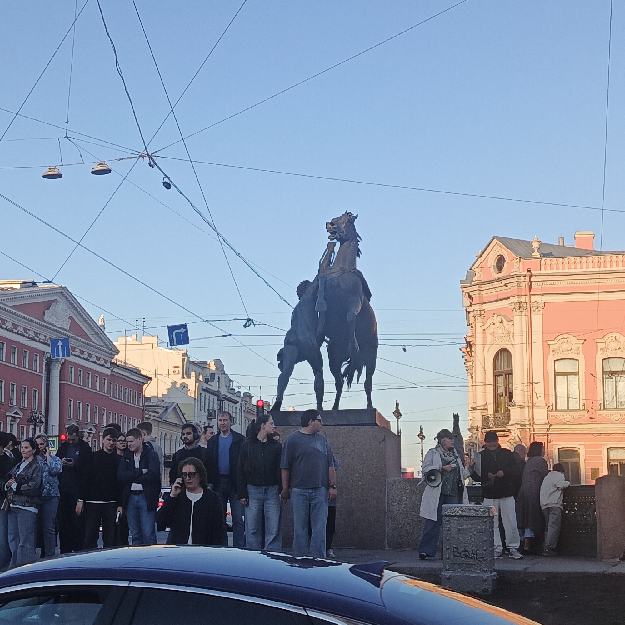 The image captures a scene in Saint Petersburg, Russia, featuring the Anichkov Bridge with the famous horse sculptures by Peter Klodt von Jürgensburg. A group of people is gathered around the bridge, likely observing or taking pictures of the sculptures. The sculptures depict a man struggling with a rearing horse. The architecture of the surrounding buildings is visible, including a pink building on the right and a red building to the left, along with the clear blue sky in the background. The presence of overhead wires and street signs adds to the urban setting. A car is visible in the foreground. Some of the people are looking at their phones or talking to each other.