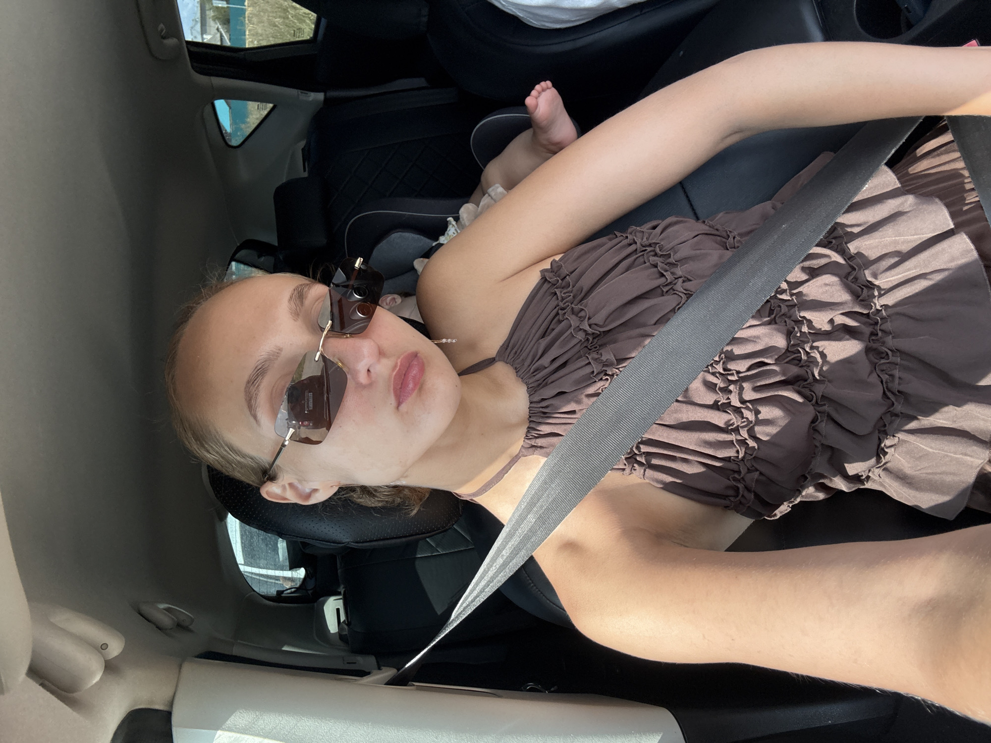 In Pirogovo, Russia, a woman with blonde hair pulled back is seated in the driver's seat of a car, looking towards the camera. She is wearing oversized sunglasses with a gold frame and a brown, ruffled halter top. Her right arm is extended, and a silver seatbelt is diagonally across her chest. In the background, a baby is visible in a car seat behind her, with its feet showing. The interior of the car is dark, with black leather seats. Outside the car window, a grassy field and buildings can be seen under a bright sky.