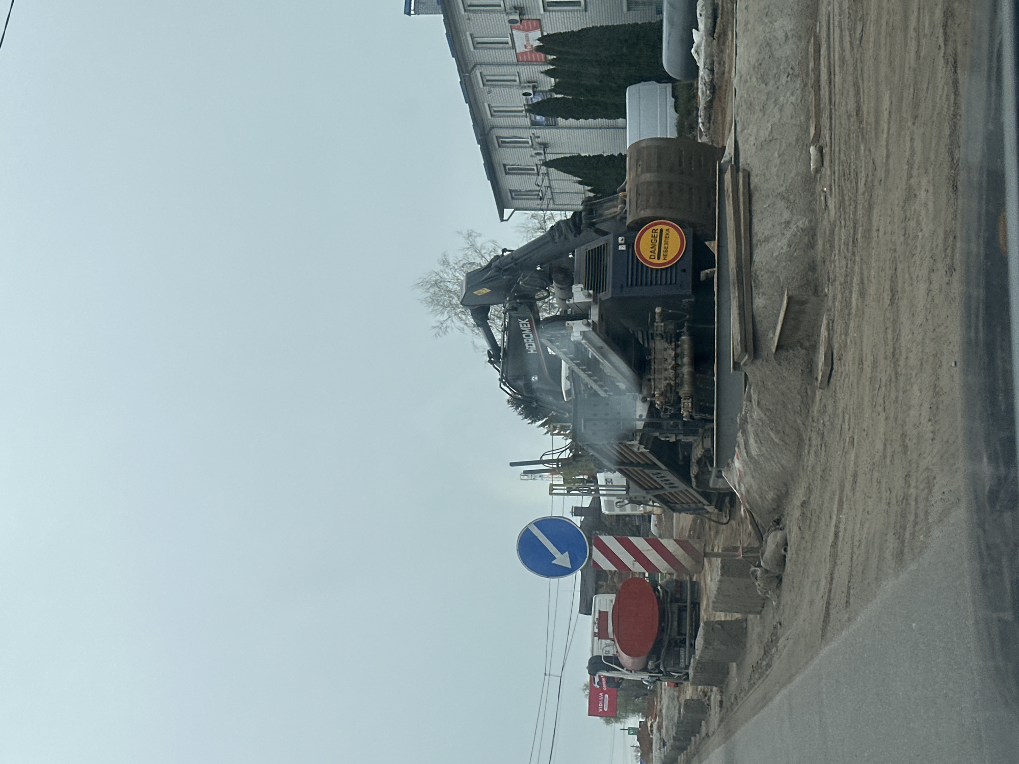The image depicts a construction site in Kyiv, Ukraine. A large Hidromek crane is positioned on the side of a road undergoing repair. A building is in the background. Signage indicates road diversion. Several construction vehicles and materials are at the site. The sky is overcast.