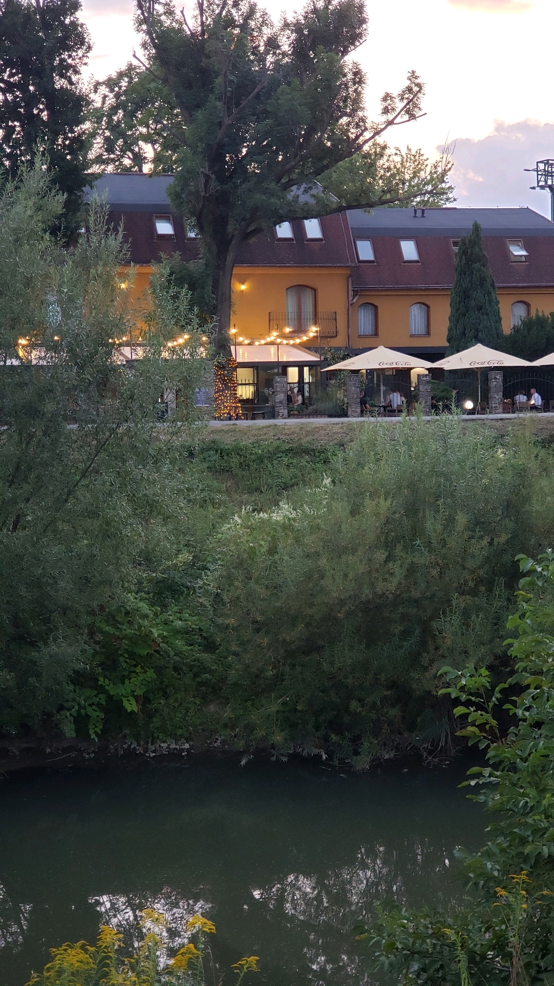A waterside building with an orange facade and a dark red roof is visible in the background. The building has several dormer windows and a balcony. String lights are draped across the front of the building, illuminating a patio area with umbrellas and outdoor seating. Several people are seated at tables on the patio. In the foreground, a body of water reflects the sky and trees. Lush green foliage borders the water and surrounds the building. The location is likely a restaurant or hotel situated on a river or lake in Prešov, Slovakia.