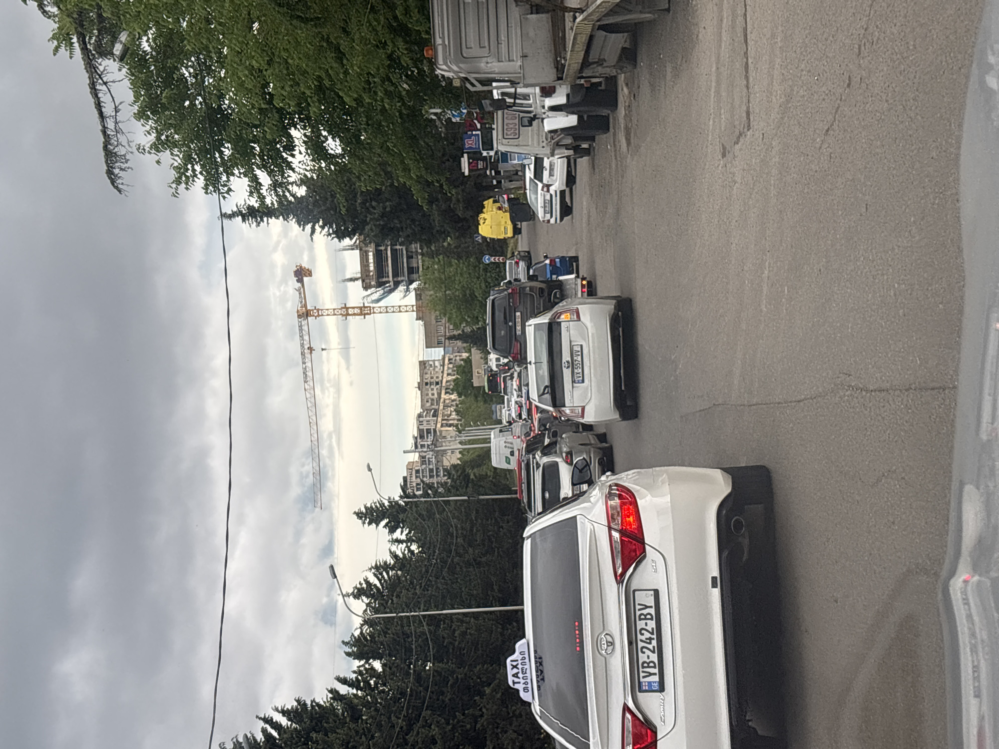 The image shows a traffic jam in Tbilisi, Georgia. The vehicles are mainly cars and trucks, with a white Toyota Camry taxi in the foreground. A construction crane is visible in the distance, indicating urban development. The street is lined with trees and buildings, and the sky is overcast. The license plate of the taxi is YB-242-BY.