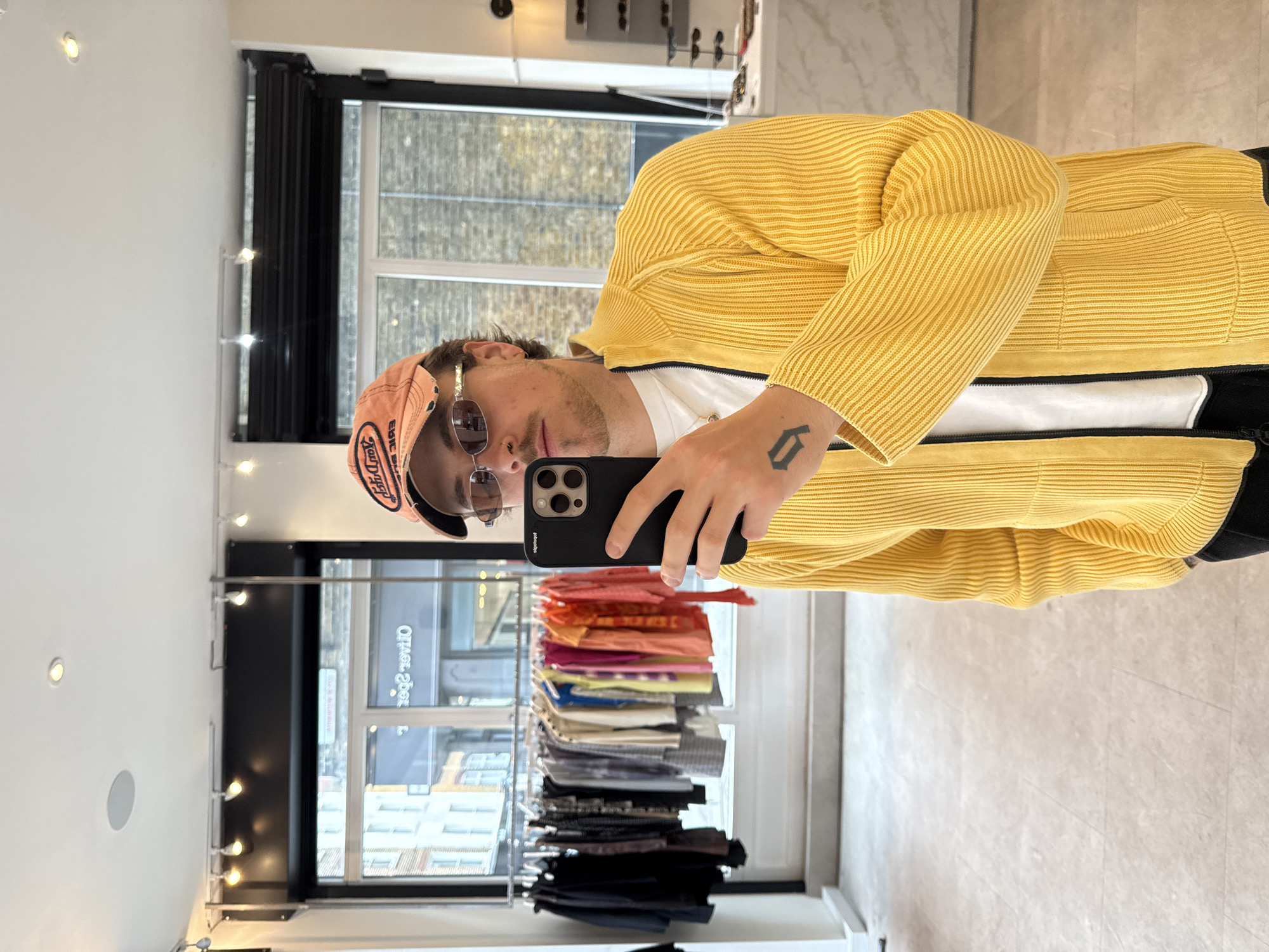 A man in a peach baseball cap, sunglasses, a white t-shirt, and a yellow knitted sweater is taking a selfie in front of a rack of clothing inside a clothing store. The man is holding a black smartphone in his right hand. The rack of clothes contains items in shades of orange, pink, purple, blue, grey, and black. The reflection in a window behind the rack of clothing reveals the store's address: 2 George Street, London. The setting appears to be a retail environment with clothing on display and a reflective window showcasing the street view.