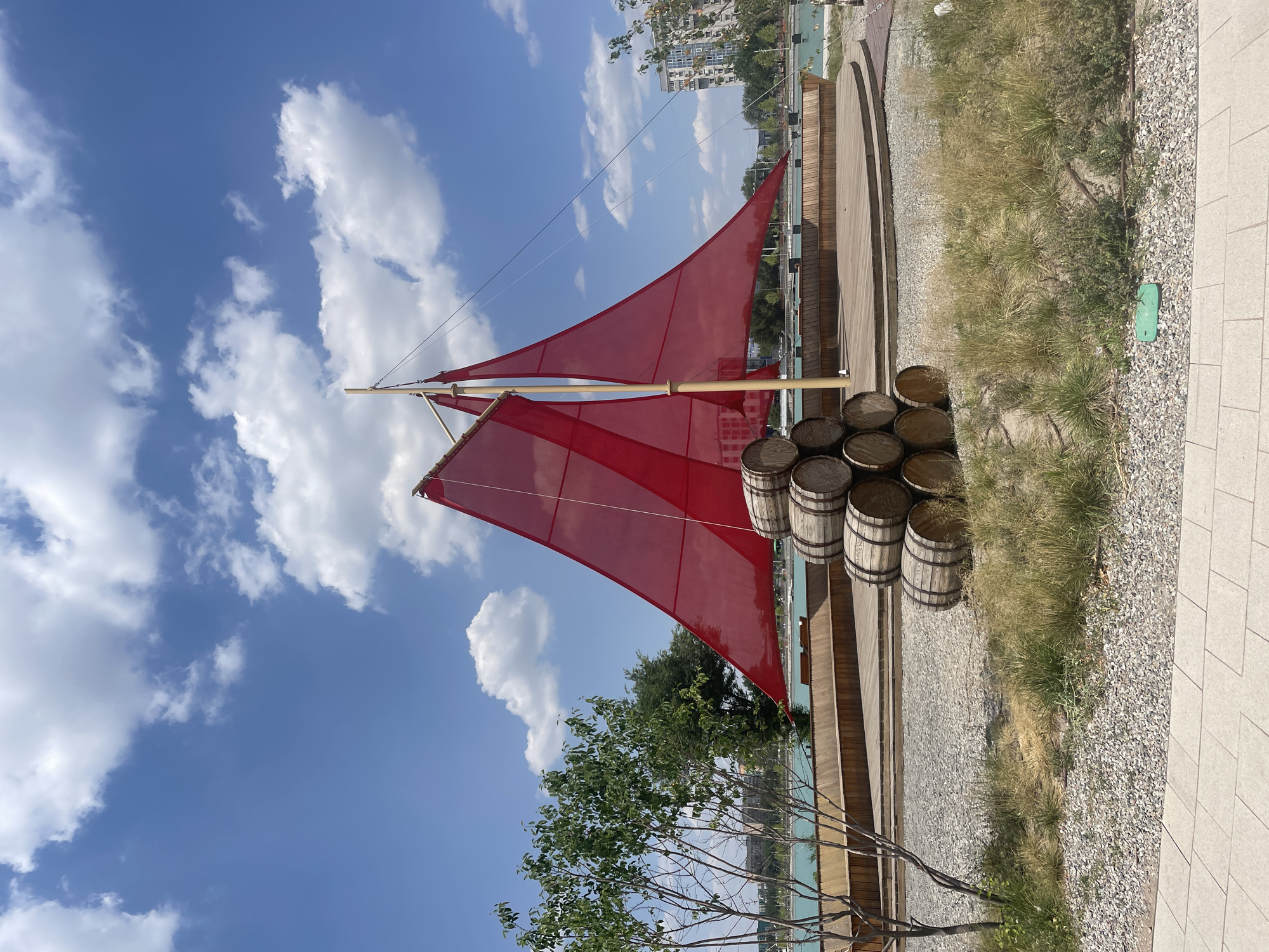 The image depicts an outdoor scene with a decorative sailboat structure in the foreground. The sailboat features two large, vibrant red sails stretched between wooden poles, resembling a classic sailing vessel. Stacked wooden barrels are placed on a wooden deck next to the sailboat.
The setting appears to be a public space, possibly a park or waterfront area, with a wooden boardwalk curving into the distance. Beyond the boardwalk, there is a body of water, likely a canal or lake, with buildings visible on the far side. The ground is covered with gravel and some dry grass. The sky is a clear blue, dotted with fluffy white clouds, suggesting a pleasant, sunny day.
There are no people visible in the image. The scene is static, focusing on the architectural elements and natural surroundings. The time of day is daytime, as indicated by the bright sunlight and clear sky. The location is identified as Almaty, Kazakhstan, though no specific landmarks are immediately apparent within the frame. The overall mood is serene and picturesque. No visible text is present in the image.