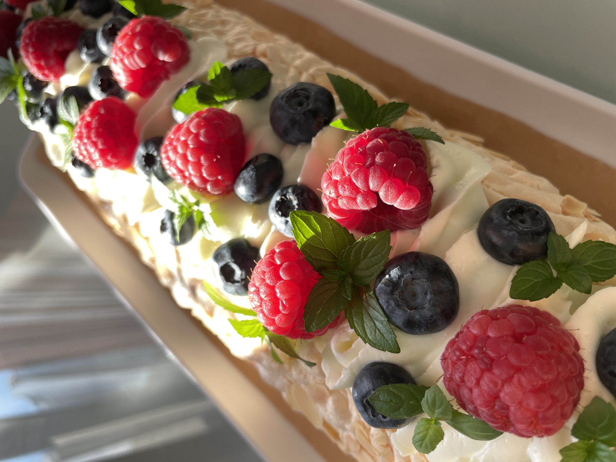 The image displays a close-up, eye-level view of a roulade cake adorned with fresh raspberries, blueberries, and mint leaves, all resting on a bed of whipped cream. The cake appears to be served in a white rectangular dish, which is placed on a light brown surface. The background is blurred and shows hints of sheer white curtains illuminated by natural light. There are no people or animals visible in the image. The scene is a still life composition focused on the dessert. The provided image does not contain any information that would allow for the identification of a specific location in the city of Saint Petersburg, Russia.