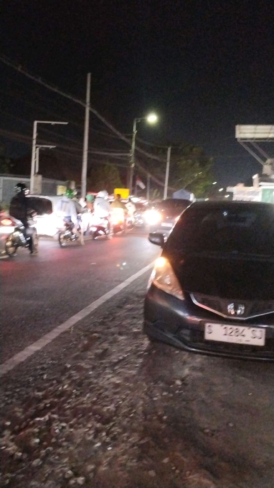 The image depicts a nighttime street scene in Canggu, Indonesia, characterized by heavy traffic and poor lighting conditions. A dark-colored car, identified by the license plate "S 1284 SJ", is prominently positioned in the foreground on the right side of the frame. Its headlights and taillights are illuminated, indicating it is either stationary or moving slowly.
On the left side of the road, several motorcycles are visible, some with riders wearing helmets. One motorcycle in the mid-ground has a passenger wearing a white shirt with blue accents, and another rider in the background is wearing a bright yellow shirt. The motorcycles appear to be part of a traffic jam or are moving in congested conditions.
The environment is urban, with streetlights casting a dim, yellow glow. Power lines and poles are visible in the background, typical of infrastructure in Indonesian cities. The road surface is asphalt, with some gravel and debris on the shoulder where the car is parked or stopped. The overall atmosphere suggests a busy, active street at night, possibly a main thoroughfare in Canggu. The image quality is low, with significant noise and blur, likely due to low light and the camera's resolution.