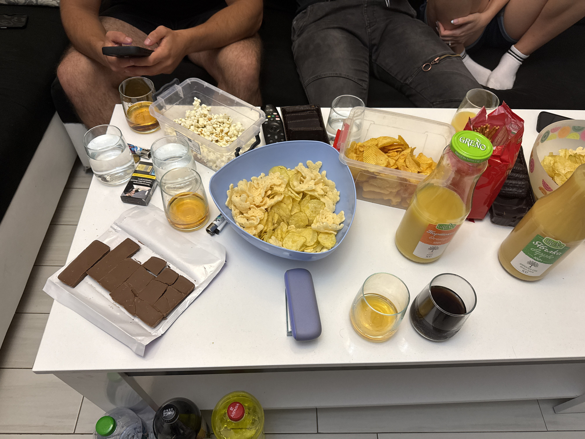 An indoor scene depicts a casual gathering with three partially visible individuals relaxing on a dark sofa, seated around a white coffee table laden with snacks and drinks. The person on the left is holding a smartphone, while another in the middle wears dark jeans, and a third on the right is seen wearing shorts and white socks with black stripes.
The table is covered with various refreshments: a clear container of popcorn, a container of potato chips, and a blue bowl filled with a mix of ruffled and puffed chips. A red bag of pretzels is also visible. For sweets, there's a partially consumed chocolate bar broken into pieces on a white wrapper.
Drinks include multiple glasses of water, two glasses containing an amber liquid, and one glass with a dark beverage. Two "GREÑO" brand juice bottles, one labeled "Портокал Orange 100%" and the other "Ябълка Apple 100% ДИРЕКТЕН СОК direct juice 1Le", are present, indicating the location is likely Bulgaria. Other beverages include a "Coca-Cola ZERO" bottle, a plain water bottle, and a bottle with a red cap labeled "ASPASIA".
Miscellaneous items on the table include a TV remote control, a pack of "Marlboro" cigarettes, a lighter, and a purple vape device or case. The overall scene suggests a relaxed social event or an evening of entertainment at home.