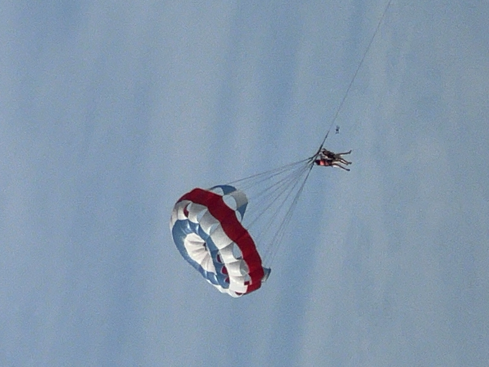In the sky above Da Nang, Vietnam, two individuals are parasailing, suspended by a large, red, white, and blue parachute. The parachute is inflated and catching the wind, with its characteristic circular shape visible. The two people, wearing life vests, are attached to the parachute by lines, and are positioned one behind the other. The backdrop is a clear, light blue sky. The scene suggests a recreational activity taking place over or near the city's coastline.