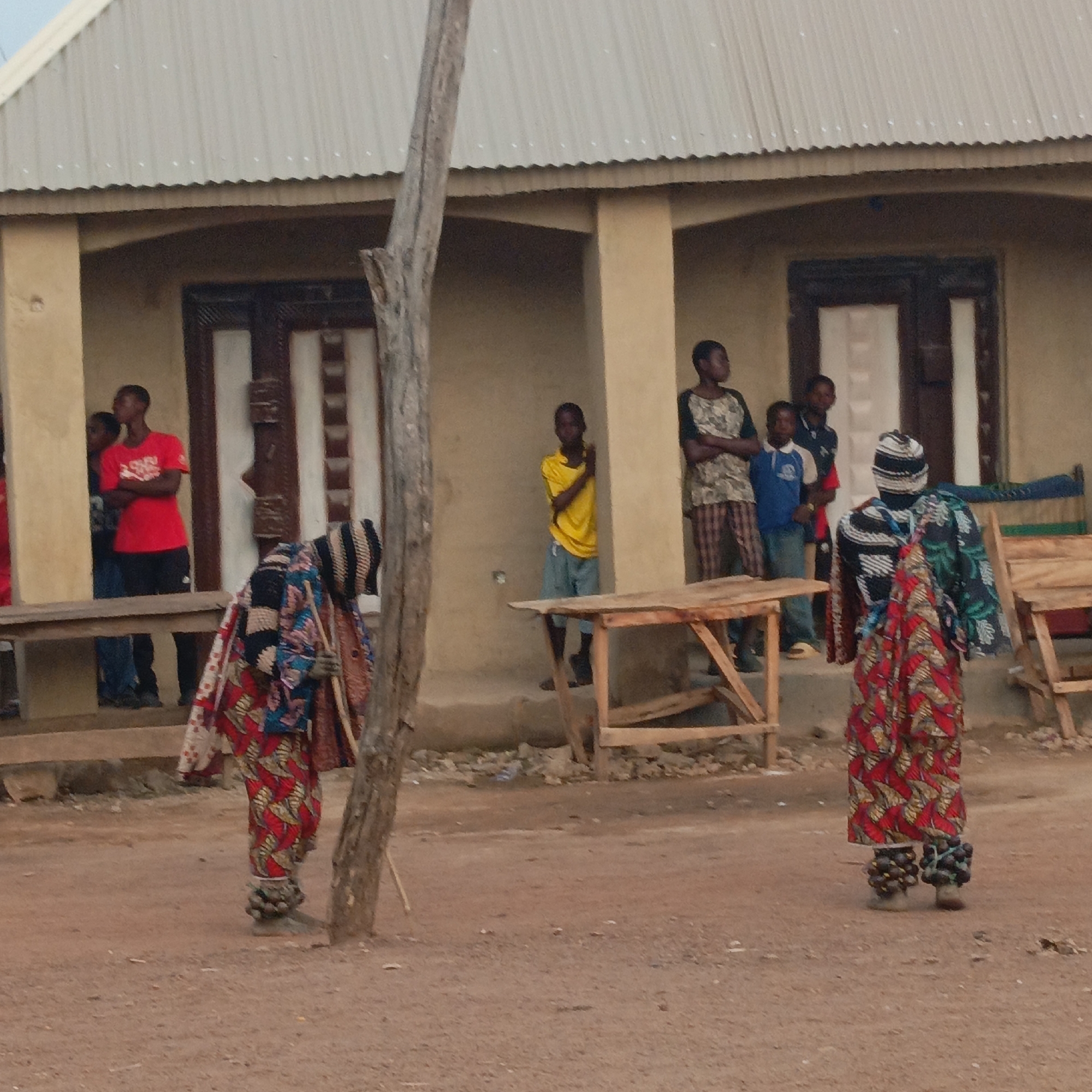 The image shows a scene in Akwanga, Nigeria. In the foreground are two individuals wearing traditional costumes, including patterned robes and beaded anklets. One is walking away from the viewer. Behind them, a group of people are standing near a building with a corrugated metal roof. There are wooden tables nearby. The ground is dirt.