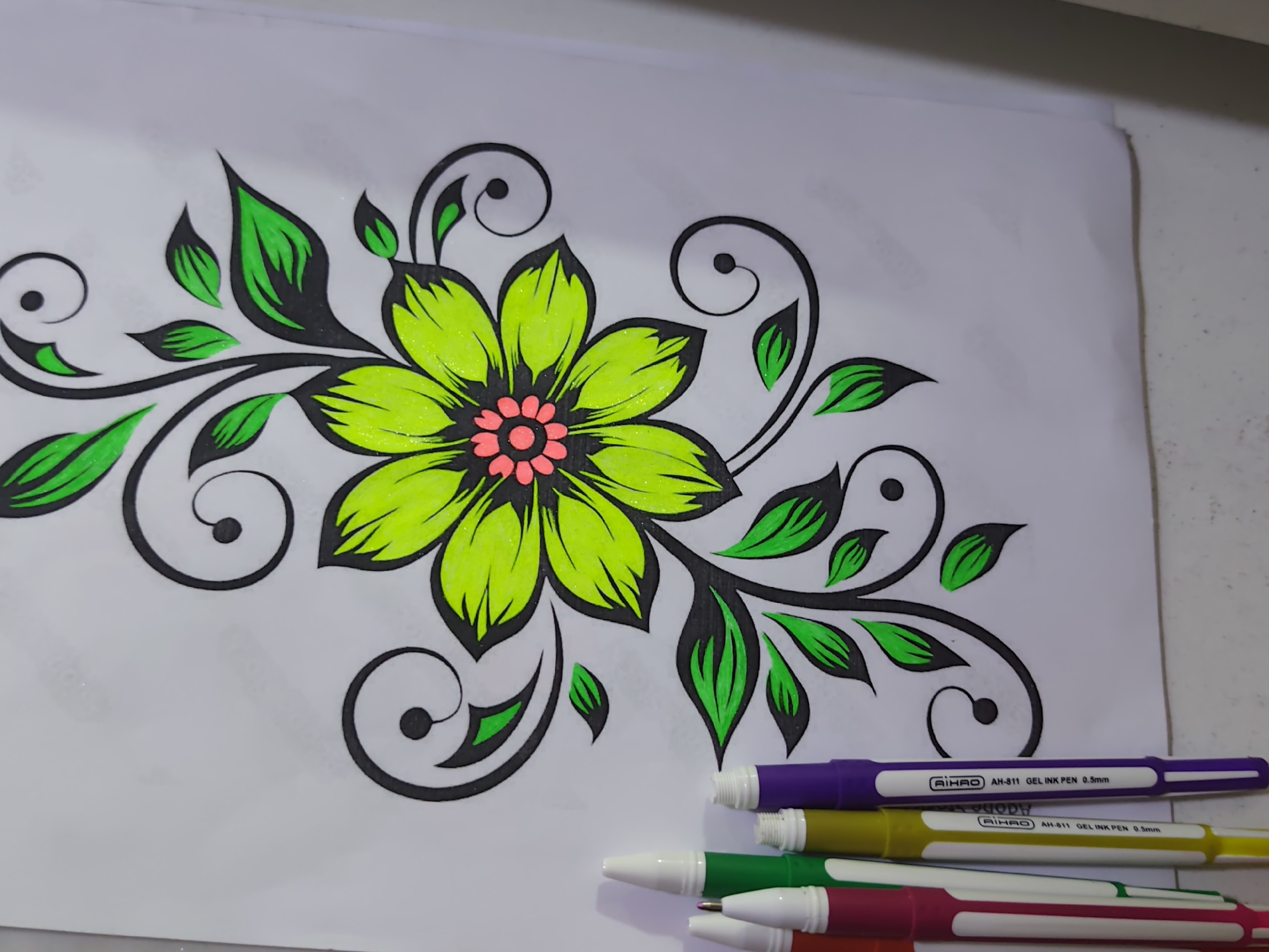 The image shows a drawing of a stylized flower with green petals outlined in black, and a pink center. The flower is surrounded by black swirls and green leaves. The drawing is on a white sheet of paper, and in the bottom right corner, there are four gel pens with purple, yellow, green, and pink ink. The drawing appears to be a piece of art or a decorative element. The background is a light gray surface. There are no people visible in the image. The scene is likely indoors, given the lighting and the presence of drawing supplies. The image does not contain any information that would allow it to be placed in Bishkek City, Kyrgyzstan.