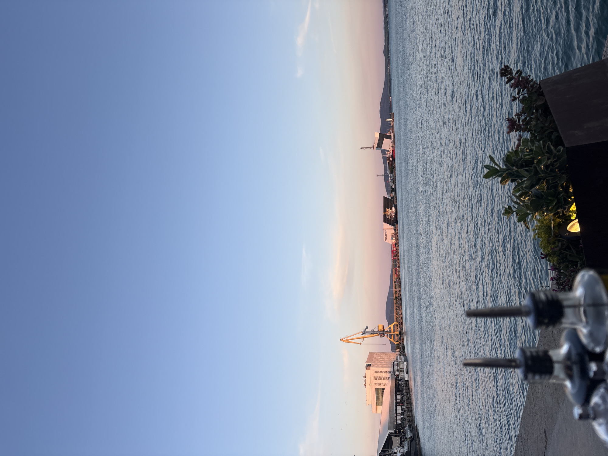 The image captures a serene view of the Burgas Bay in Burgas, Bulgaria, during what appears to be sunset or early evening. The sky is a clear, soft blue transitioning into hues of pink and orange near the horizon, suggesting the presence of some clouds. The calm, blue water of the bay occupies the lower half of the image, reflecting the gentle light.
In the middle ground, the Burgas Port is visible, featuring a mix of industrial and modern architectural elements. A prominent bright yellow crane stands tall, indicating ongoing port activities. Several buildings line the waterfront, including a large, white, modern structure with a distinctive patterned facade. Ships and boats are docked along the piers, and other port infrastructure, such as smaller cranes and buildings, can be seen in the distance. The landscape in the far background is characterized by a hilly, dark silhouette.
In the foreground, two glass bottles with metallic spouts are positioned on a stone surface, suggesting the setting might be an outdoor seating area of a restaurant or bar. A planter box with green foliage and some reddish-purple flowers is also visible in the lower right corner. A faint glow from a light fixture is noticeable within the planter. No people are explicitly visible in the image, but the presence of the restaurant setting implies a possibility of patrons.