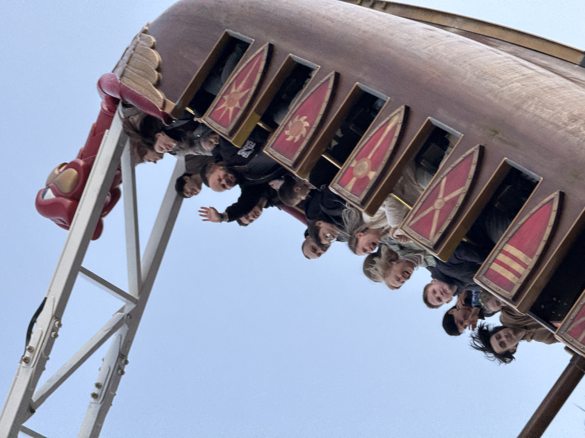 A tilted, upward shot depicts a "Viking Ship" amusement park ride at Sochi Park in Sochi, Russia. Passengers, visible through shield-decorated openings, are experiencing the thrill of the ride as it swings upward against a pale blue sky. The ride's arm, built with a lattice framework, connects to the Viking Ship, which is designed with a decorative dragon head on its bow. Passengers appear to range from children to adults and are captured in varied expressions of excitement.