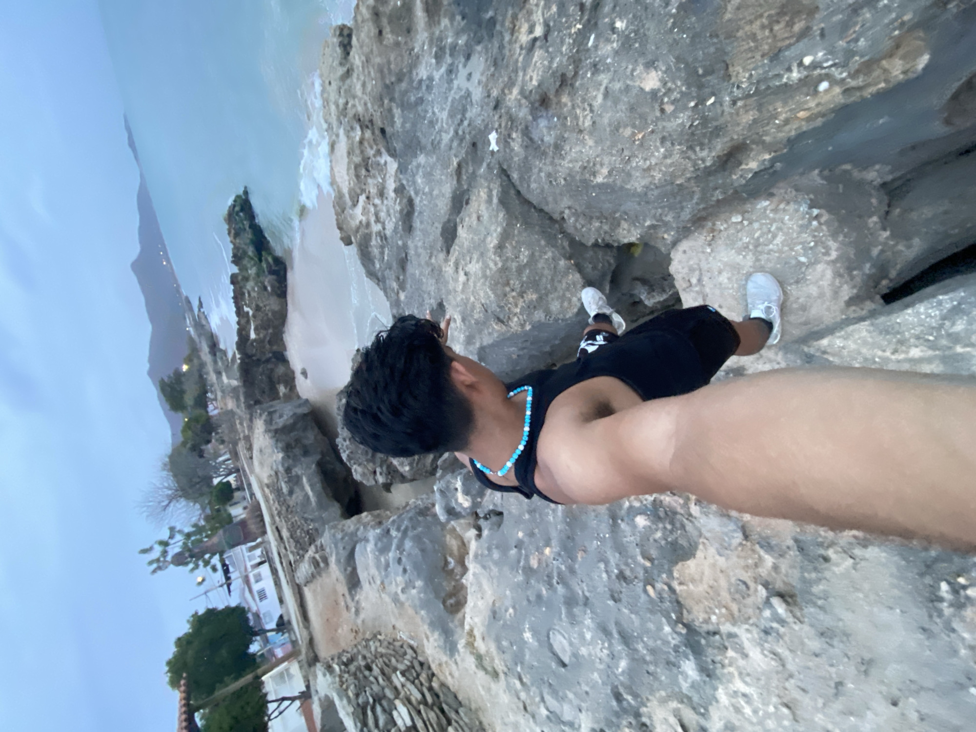 A man is positioned on rocky terrain with a view of the coastline. The water is a light turquoise color, and there is a beach visible in the distance. The terrain consists of gray rocks with some white elements. The man is wearing black shorts, a black tank top, white shoes, and a turquoise bead necklace. Buildings and greenery are visible along the coastline. This is the city of Juan Griego, Venezuela.