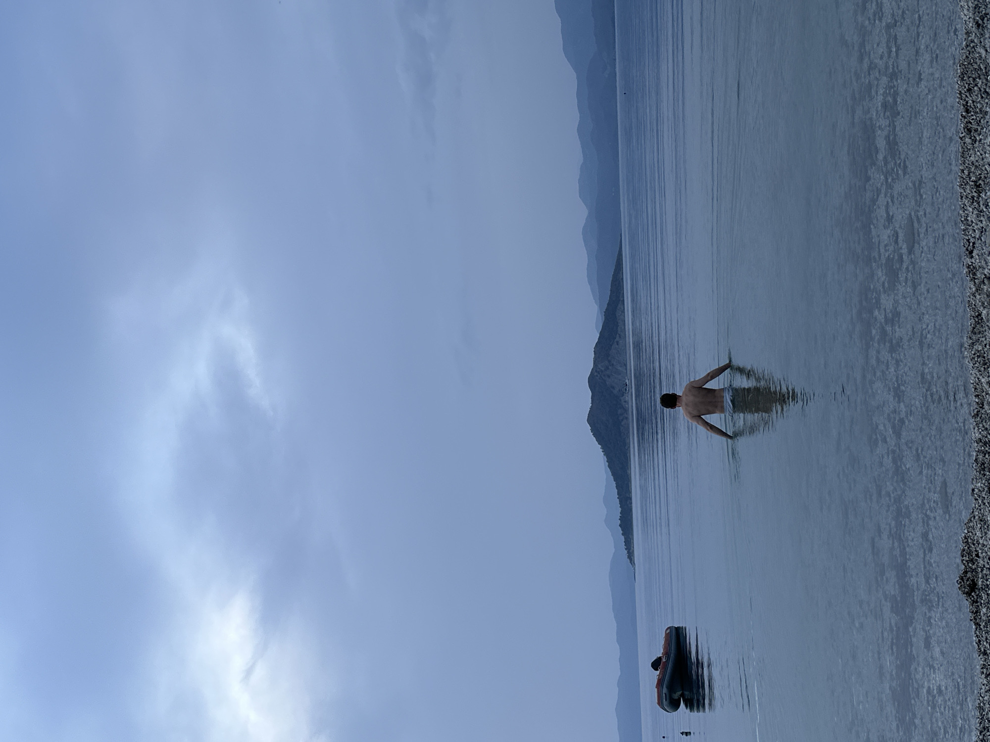 The image depicts a serene waterscape under an overcast sky. A lone individual, appearing to be a man, stands waist-deep in calm, bluish-grey water, facing away from the camera. His upper torso is visible above the water, with his arms slightly outstretched. Further to the left in the water, a dark-colored inflatable boat, possibly containing a person, floats gently.
In the background, a range of mountains stretches across the horizon, rendered in muted tones of blue and grey, suggesting distance and atmospheric haze. The sky above is uniformly overcast, with soft, diffused light, indicating a cloudy day or a time around dawn or dusk. To the right of the frame, a pebbly or rocky shoreline is visible, bordering the clear water.
The scene conveys a sense of tranquility and peacefulness. There is no visible text in the image. The setting of mountains, calm water, and a pebbly shore is consistent with natural landscapes found in locations like Fethiye, Turkey, which features lakes and sheltered sea bays.