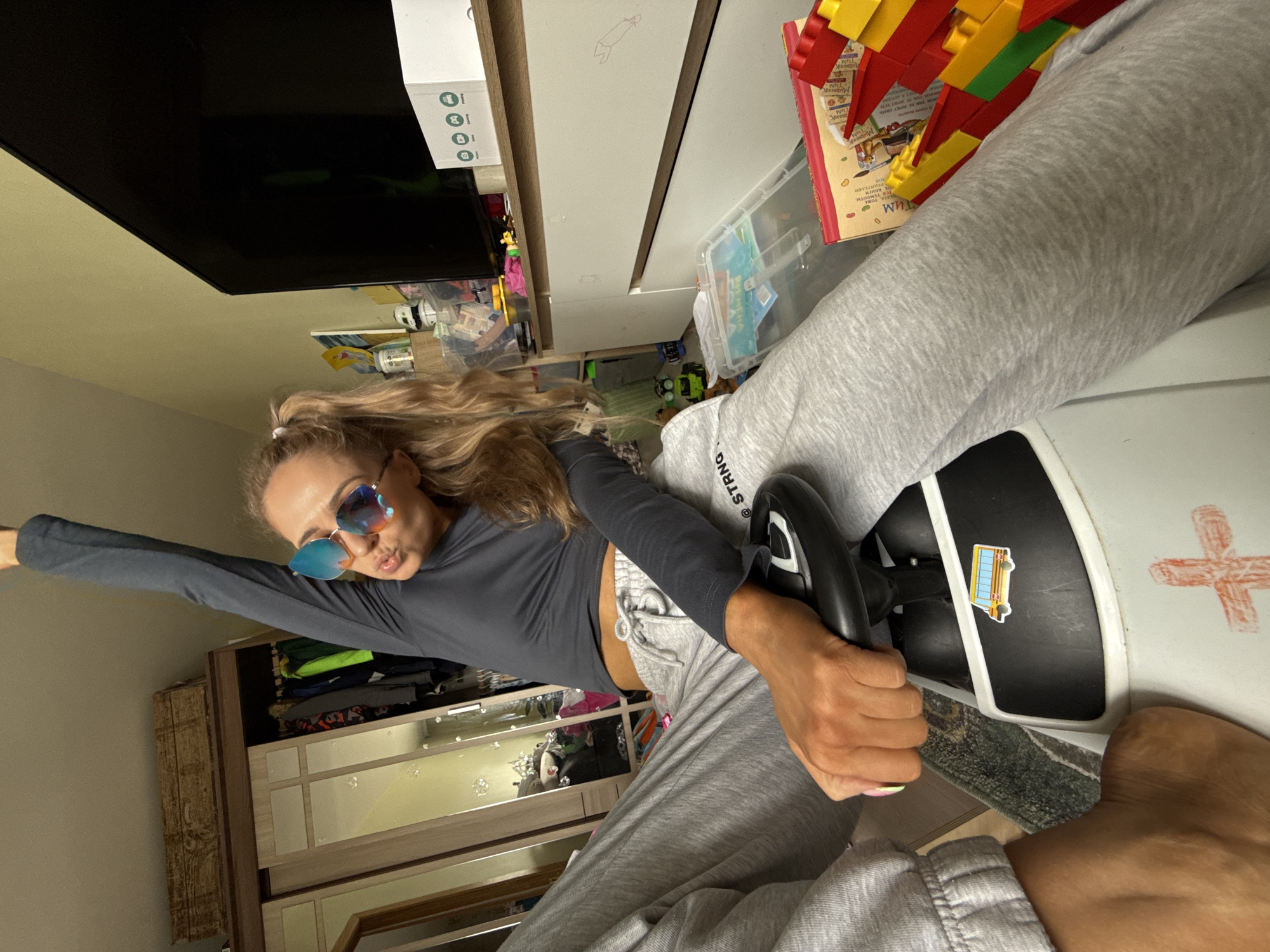A young woman with long, wavy blonde hair and blue mirrored sunglasses is captured in a playful pose indoors in Chaikovsky, Russia. The image is oriented sideways, giving a dynamic, tilted perspective of the room. She is wearing a dark grey long-sleeved crop top and light grey sweatpants, one leg extended and barefoot.
The woman is seated in or on a white ride-on toy car, gripping its black steering wheel with both hands and striking a pose with her left arm outstretched. A yellow school bus sticker is visible on the toy car's dashboard, and a hand-drawn red cross is on its side. She has a fun, puckered expression on her face.
The background reveals a casual home environment. To her left (top in the rotated image), a large black flat-screen television is mounted above a white box displaying four green circular icons. To her right, a light-colored cabinet or dresser holds a clear plastic bin filled with colorful building blocks and a book with a red and yellow cover, featuring Cyrillic text including "Малышам" (for toddlers). Below the television and cabinet, various toys and household items are scattered, contributing to a lived-in atmosphere. Another wooden cabinet with reflective panels is visible further to the left. Partially visible text on her sweatpants reads "STRNG". The overall scene suggests a lighthearted moment within a family home.