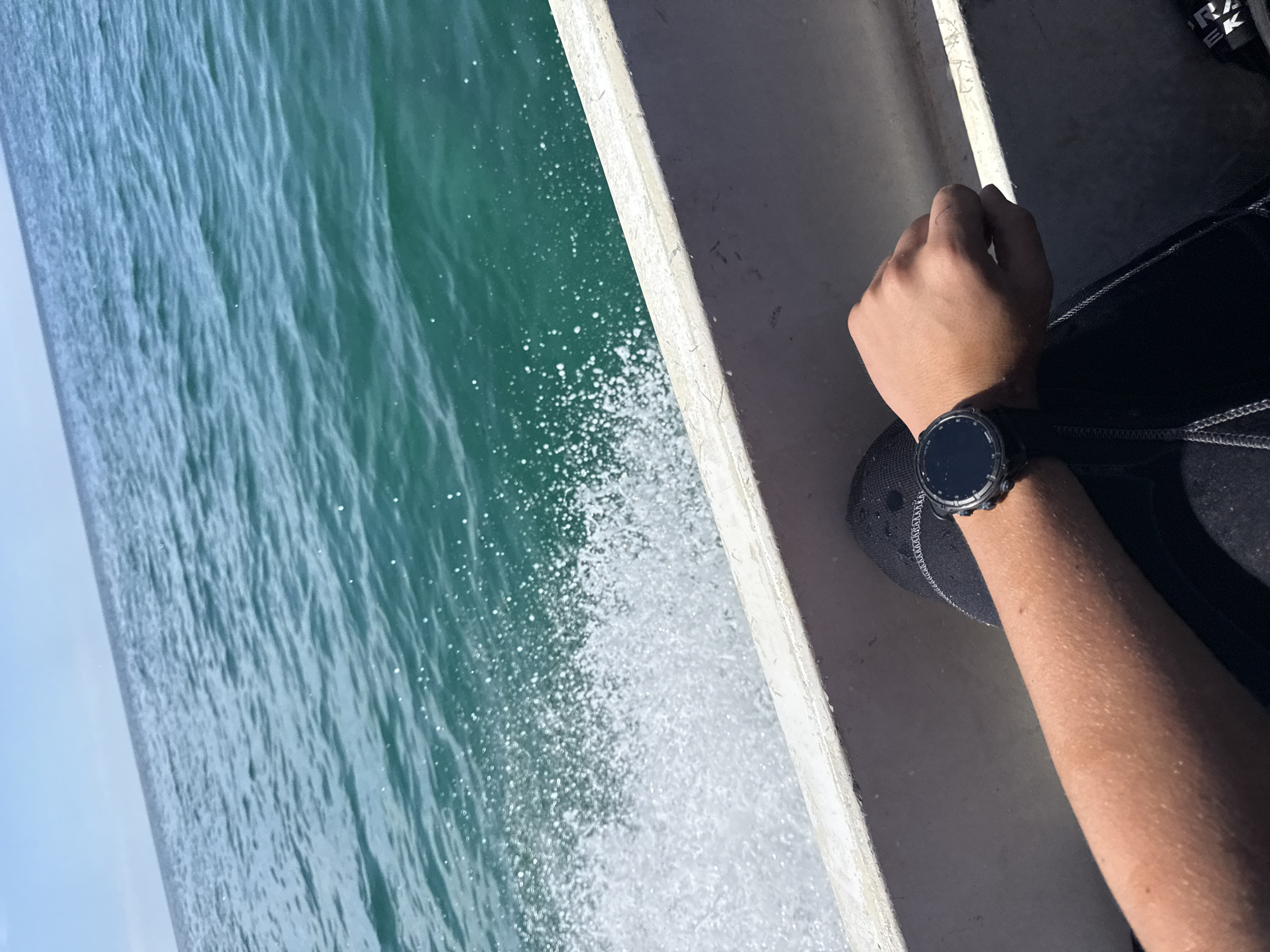 An arm wearing a black smartwatch rests on the side of a white boat. The boat is moving through clear, turquoise water, creating a spray of white foam. The sky above is a pale blue. The scene appears to be a daytime view from a boat on the sea, possibly near Limenaria, Greece, given the clarity of the water and the bright sunlight.