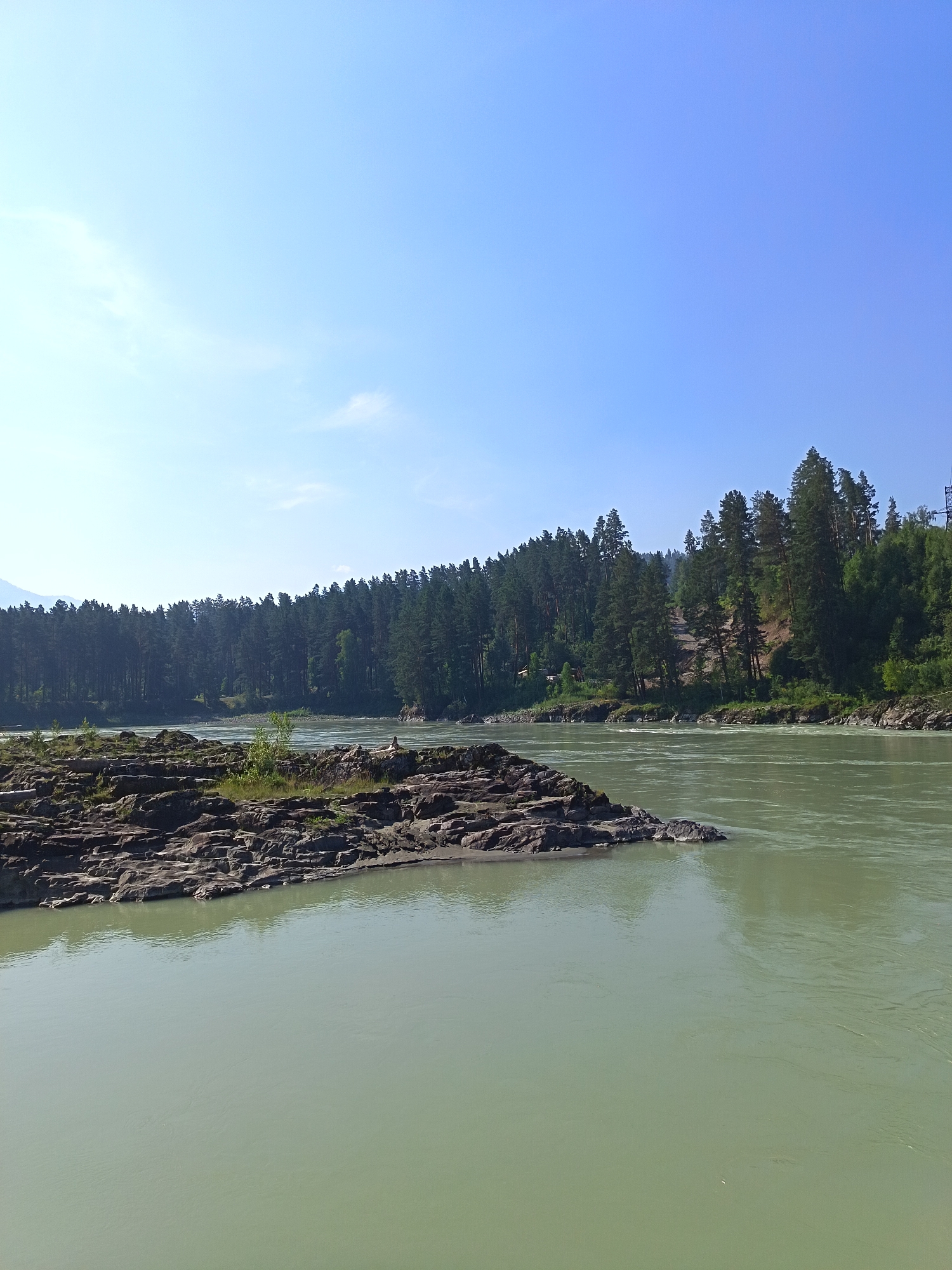 The image depicts a wide, calm river flowing through a forested landscape under a clear blue sky. In the foreground, a cluster of dark, rugged rocks emerges from the water, with some sparse green vegetation growing on their surface. The river's water is a muted, greenish-blue color, reflecting the sky and surrounding trees.
The middle ground is dominated by a dense forest of tall, dark green pine trees that line the riverbank. The trees are tightly packed, forming an unbroken wall of foliage that stretches across the horizon. The terrain behind the trees appears to rise, with some lighter patches suggesting slopes or clearings.
In the far distance, the sky is a brilliant, cloudless blue, with a faint haziness near the horizon. There are no visible people or animals in the image, nor any apparent man-made structures apart from what might be a distant, small building nestled among the trees on the left side of the image, and a metal pylon structure on the far right. The overall impression is one of natural tranquility and remoteness.
The image appears to have been taken from a slightly elevated viewpoint, looking across the river towards the forested bank. The lighting suggests it is daytime, likely late morning or early afternoon, given the bright illumination and clear visibility. The absence of any significant weather phenomena indicates a stable and pleasant atmospheric condition.