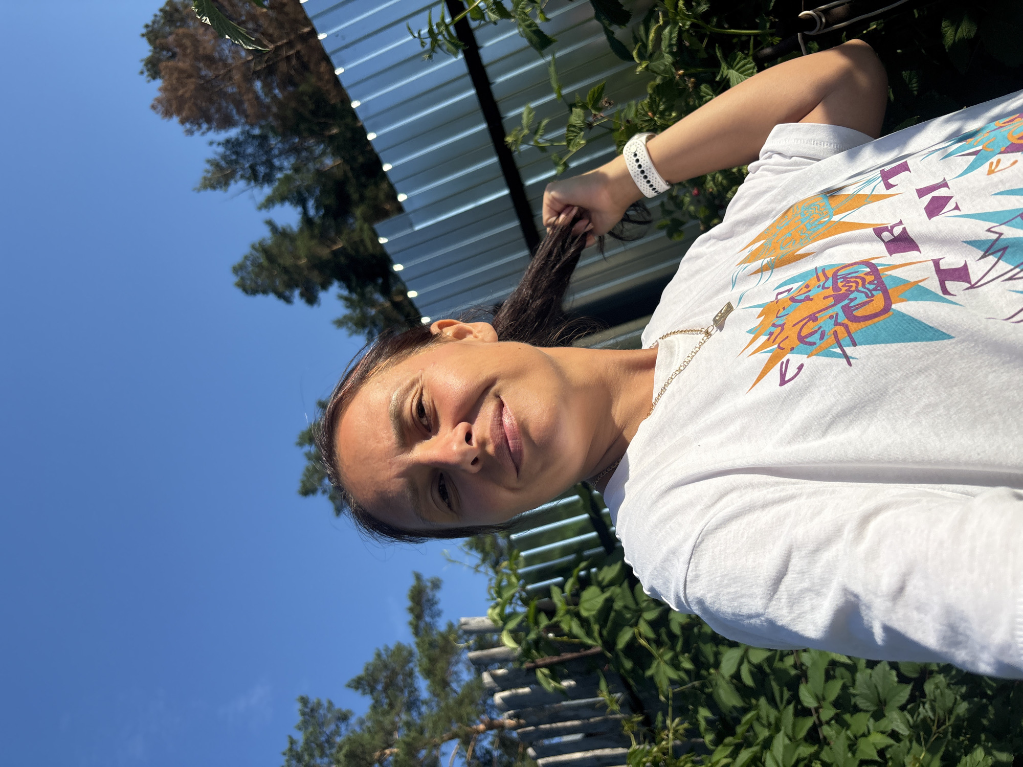 A woman is taking a selfie outdoors, in front of a background of greenery and a fence. She has dark hair pulled back and is wearing a white t-shirt with a colorful graphic on it, a gold chain necklace, and a white watch. The image appears to be taken during the daytime, with a clear blue sky visible. There are trees and a corrugated metal fence in the background. The location is a backyard in Chaikovsky, Russia.