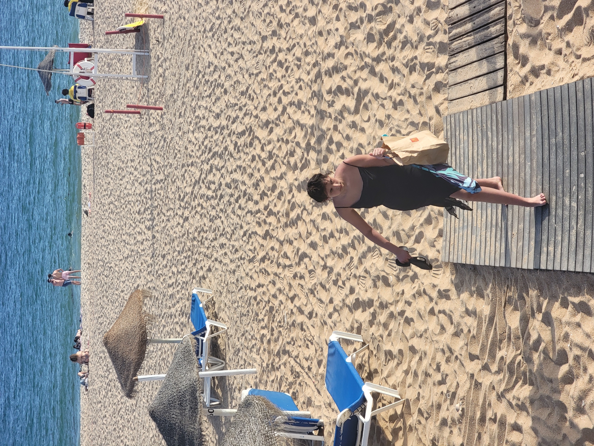 The media file depicts a sunny day at Quarteira beach, Portugal. The scene is dominated by a sandy beach with the turquoise waters of the ocean on the left. Several beach chairs with blue seats and white frames are visible, some with woven sunshades. A wooden boardwalk runs along the sand, providing a pathway.
In the foreground, a person is shown walking on the boardwalk, holding a brown paper bag with the McDonald's logo visible. They are wearing a dark dress and patterned shorts. Further down the beach, other beachgoers are seen enjoying the sun and sea. Some individuals are standing in the water, while others are relaxing on the sand or seated on chairs.
In the background, a lifeguard station is present, equipped with a lifebuoy and a red box. A rope is also attached to the station. The overall atmosphere appears relaxed and leisurely, characteristic of a typical summer day at the beach. The bright sunlight suggests it is midday.
