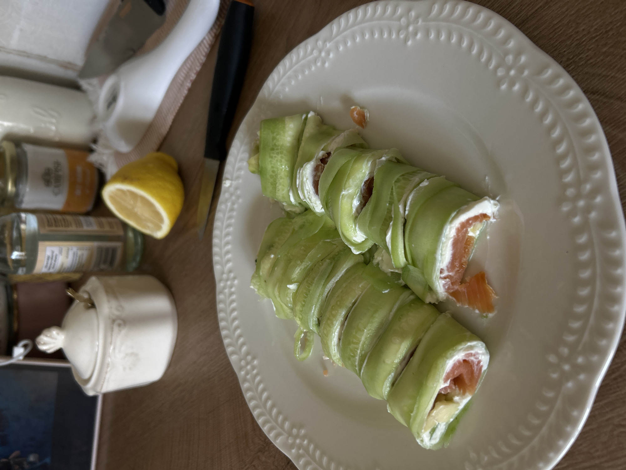 The image shows a close-up, overhead view of two cucumber rolls filled with cream cheese and salmon, arranged on a white decorative plate. The rolls are sliced into bite-sized pieces and are positioned on the right side of the plate. A fork is visible on the left side of the plate, and a knife lies behind the plate. In the background, various items are out of focus, including a lemon, a sugar bowl with a spoon, and bottles. The overall setting appears to be a domestic environment, possibly a kitchen or dining area, with a wooden table as the surface. The image does not provide any specific visual cues or context that would allow for the identification of its location within Plovdiv, Bulgaria.