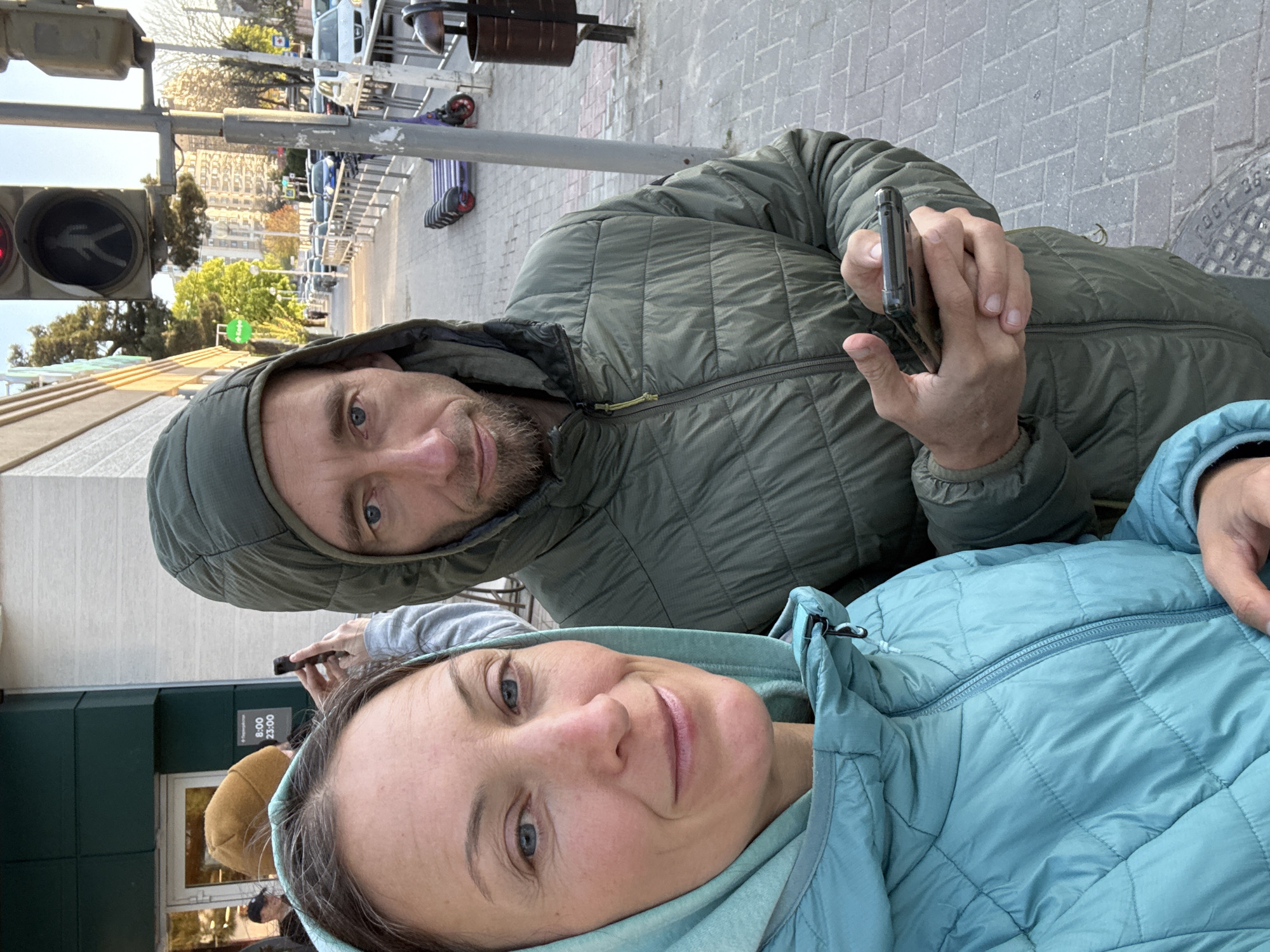 The image is a selfie depicting a man and a woman in an urban setting in Gelendzhik, Russia. The man, positioned slightly behind and to the left of the woman, has a beard, blue eyes, and wears a dark olive green hooded quilted jacket. He holds a smartphone in his right hand, likely taking the photo, and looks at the camera with a neutral expression. The woman, in the foreground, has brown hair and blue eyes, wearing a light blue hooded quilted jacket, and offers a slight smile to the camera.
The background reveals a city street scene during daytime. A pedestrian traffic light displaying a red "walking man" signal is visible on the far left. Beyond the subjects, a paved sidewalk extends, bordered by buildings and some trees. Elements like scooter parking racks and parked vehicles indicate a bustling street. The sky appears clear and bright.
Notable details include a sign on a dark green building to the lower left, displaying business hours "8:00 - 23:00." A manhole cover on the ground is partially visible, showing Russian text "ГОСТ 363," confirming the location context of Russia. Both individuals are dressed in warm jackets, suggesting cool but not severe weather. The overall atmosphere is casual, capturing a moment as the couple poses for the camera.