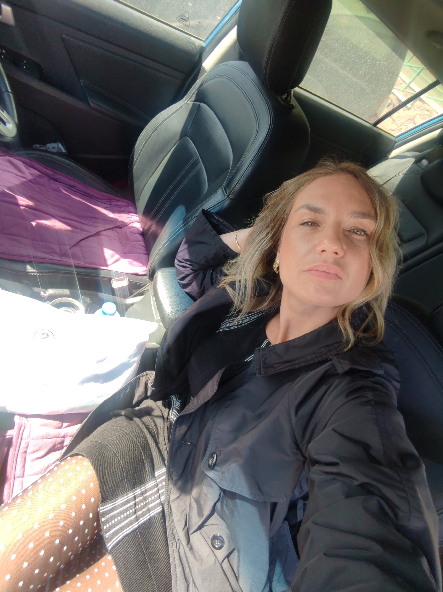 A woman with light brown hair and fair skin sits in the passenger seat of a car. She wears a black jacket, a black dress, and polka-dot patterned tights. Her left hand is behind her head. A purple blanket is on the driver's seat. The location is unknown and there is no evidence of Petropavlovsk-Kamchatsky, Russia.