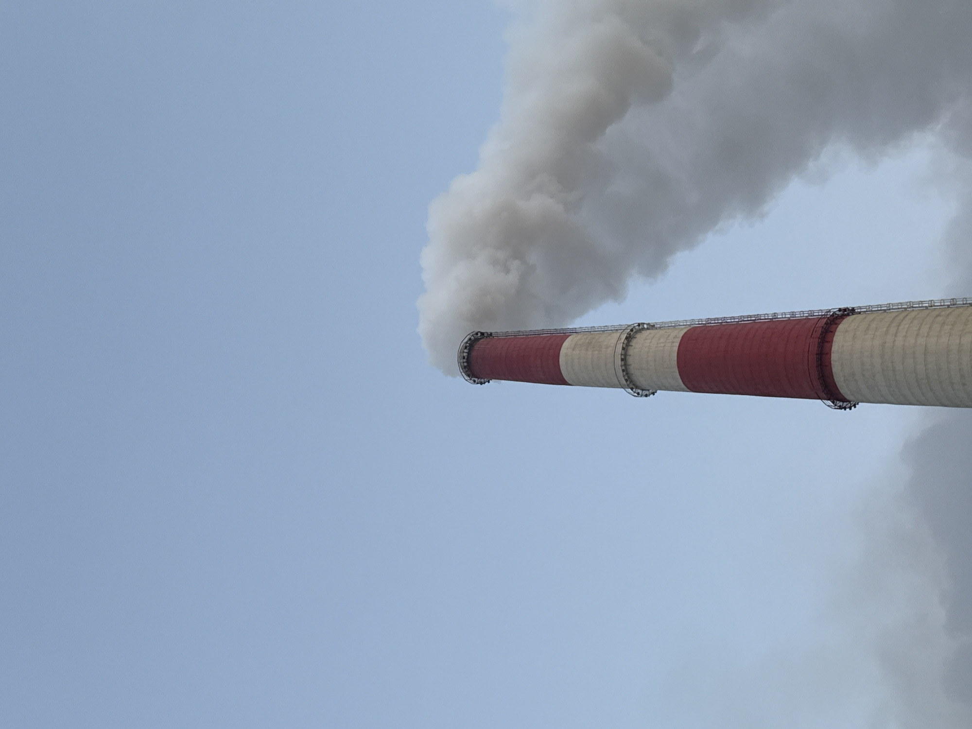An industrial chimney with red and white bands stands tall against a clear blue sky. Thick white smoke billows from the top of the chimney and drifts to the right. The chimney is a tall, cylindrical structure with visible metal supports or platforms at intervals. The image is cropped to focus on the upper portion of the chimney and the escaping smoke, with the vast expanse of the sky dominating the background. The scene appears to be an outdoor industrial setting, likely a power plant or factory, located in the city of Norilsk, Russia, known for its industrial output and resulting atmospheric emissions. There are no people or other identifiable subjects in the image.