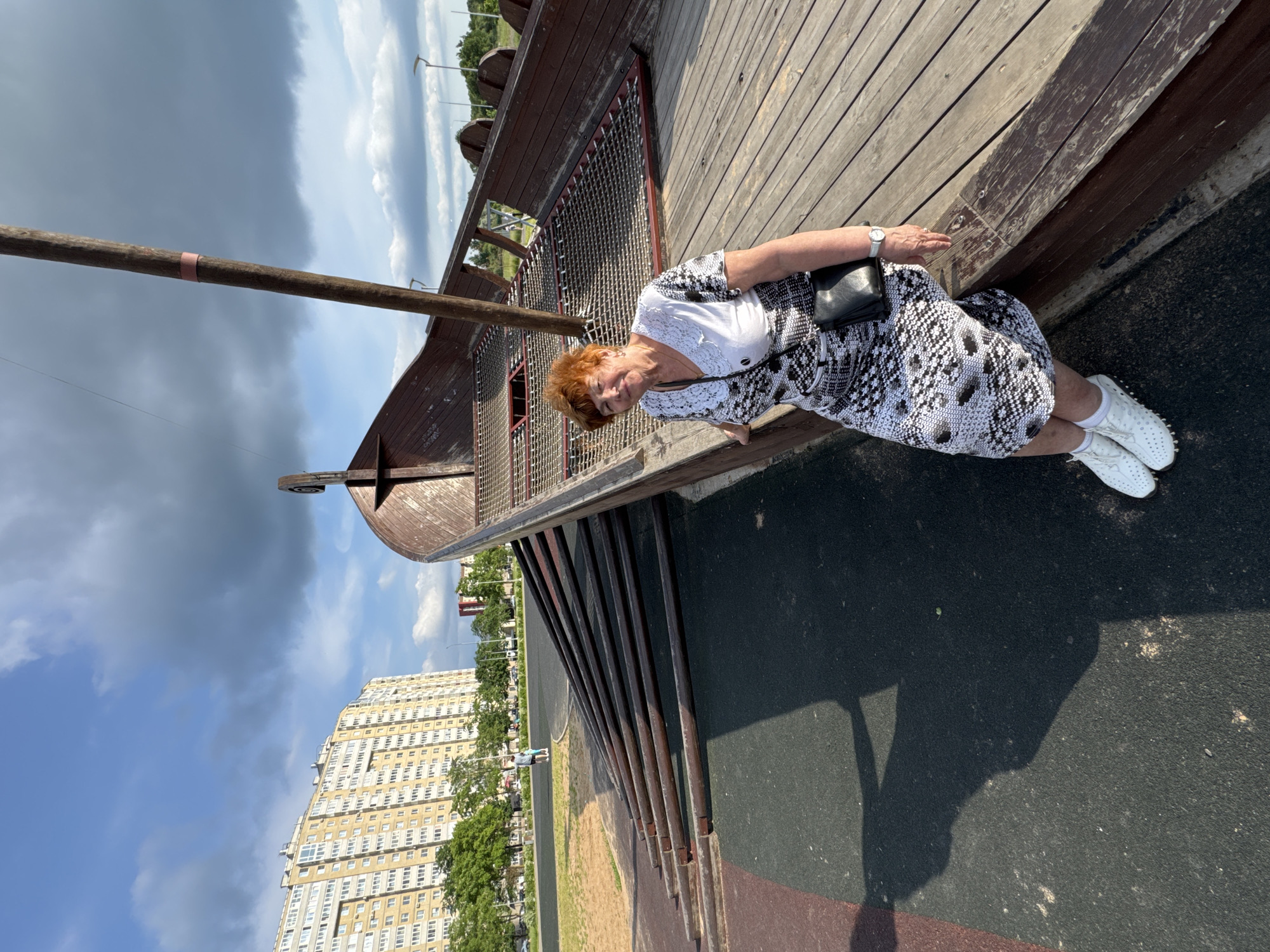 An elderly Caucasian woman with short, reddish-blonde hair and fair skin is sitting on the wooden deck of a large, ship-like structure. She is wearing a black and white patterned dress, white perforated shoes, white socks, and a silver watch. She has a black purse tucked under her arm. The woman is smiling and looking at the camera. The ship structure appears to be part of a playground or park, with a wooden hull and a mast. In the background, a tall, multi-story apartment building is visible, along with trees, a street, and a cloudy sky. The ground is a mix of dark gray and reddish-brown asphalt. The overall scene suggests a sunny day in an urban park. The location is the city of Kirishi, Russia.