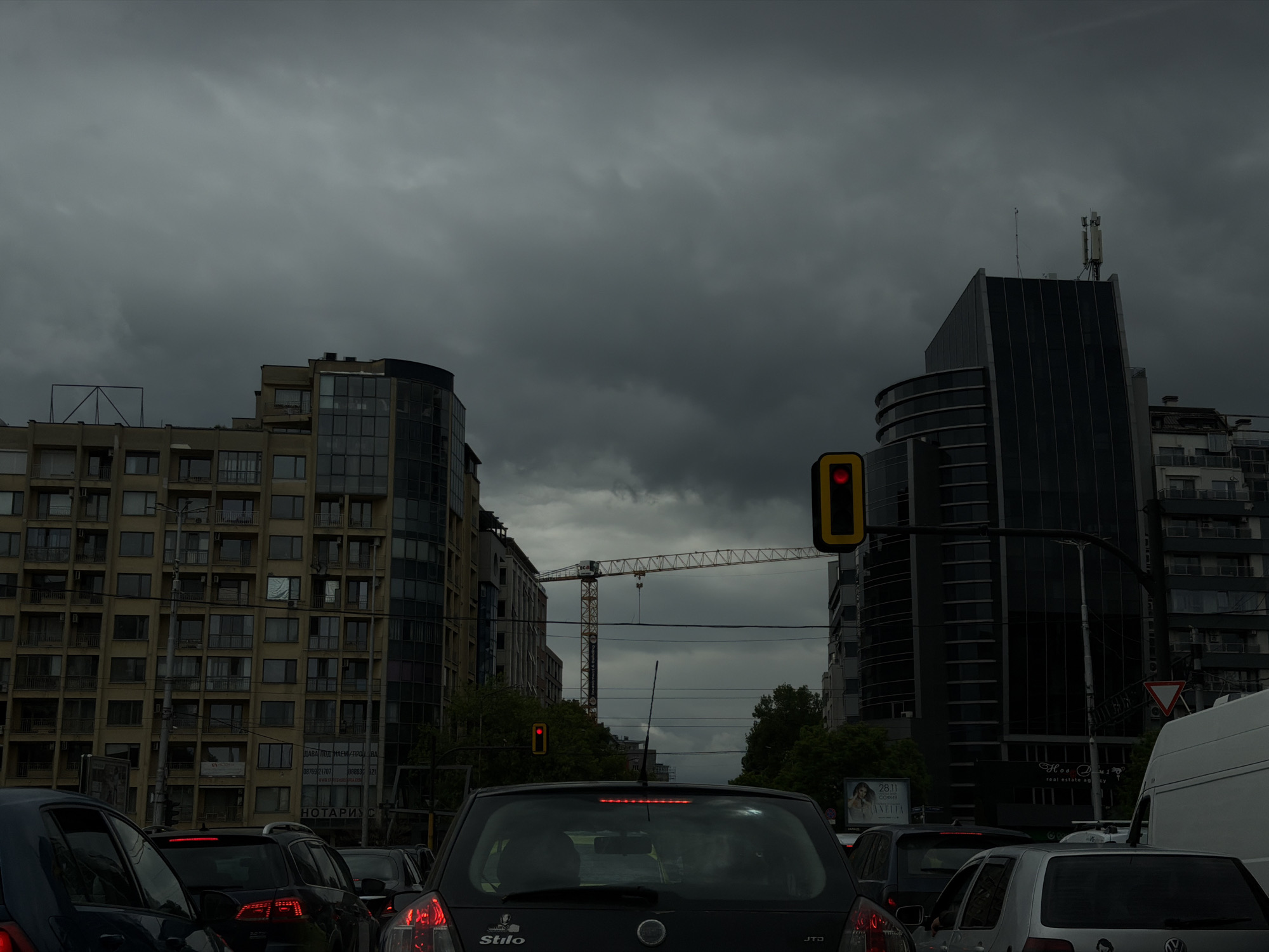 The image depicts a street scene in Sofia, Bulgaria on an overcast day. In the foreground are several cars queued at a red traffic light. Dominating the background are multi-story buildings: a yellow-beige building on the left and a dark, modern building on the right. A construction crane is visible between the buildings. The sky is dark with ominous clouds.