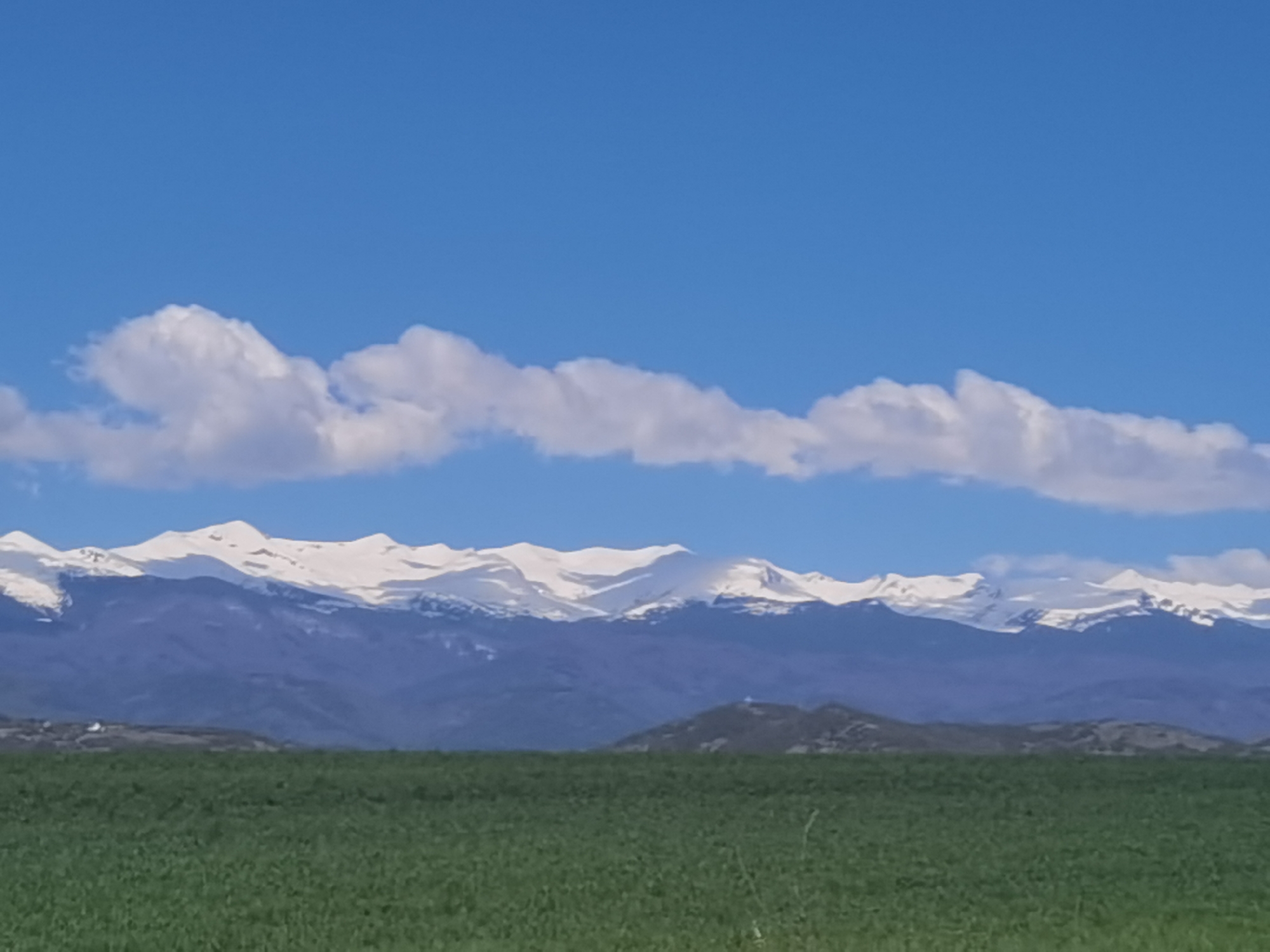 The image shows a rural landscape under a bright blue sky with scattered white clouds. In the distance, a mountain range with snow-capped peaks stretches across the horizon. Below the mountains, a range of lower hills is visible. The foreground is dominated by a green field. The location is likely near Baldevo, Bulgaria, based on the presence of the mountain range. No people, subjects, objects, actions, or interactions are visible other than the natural elements.