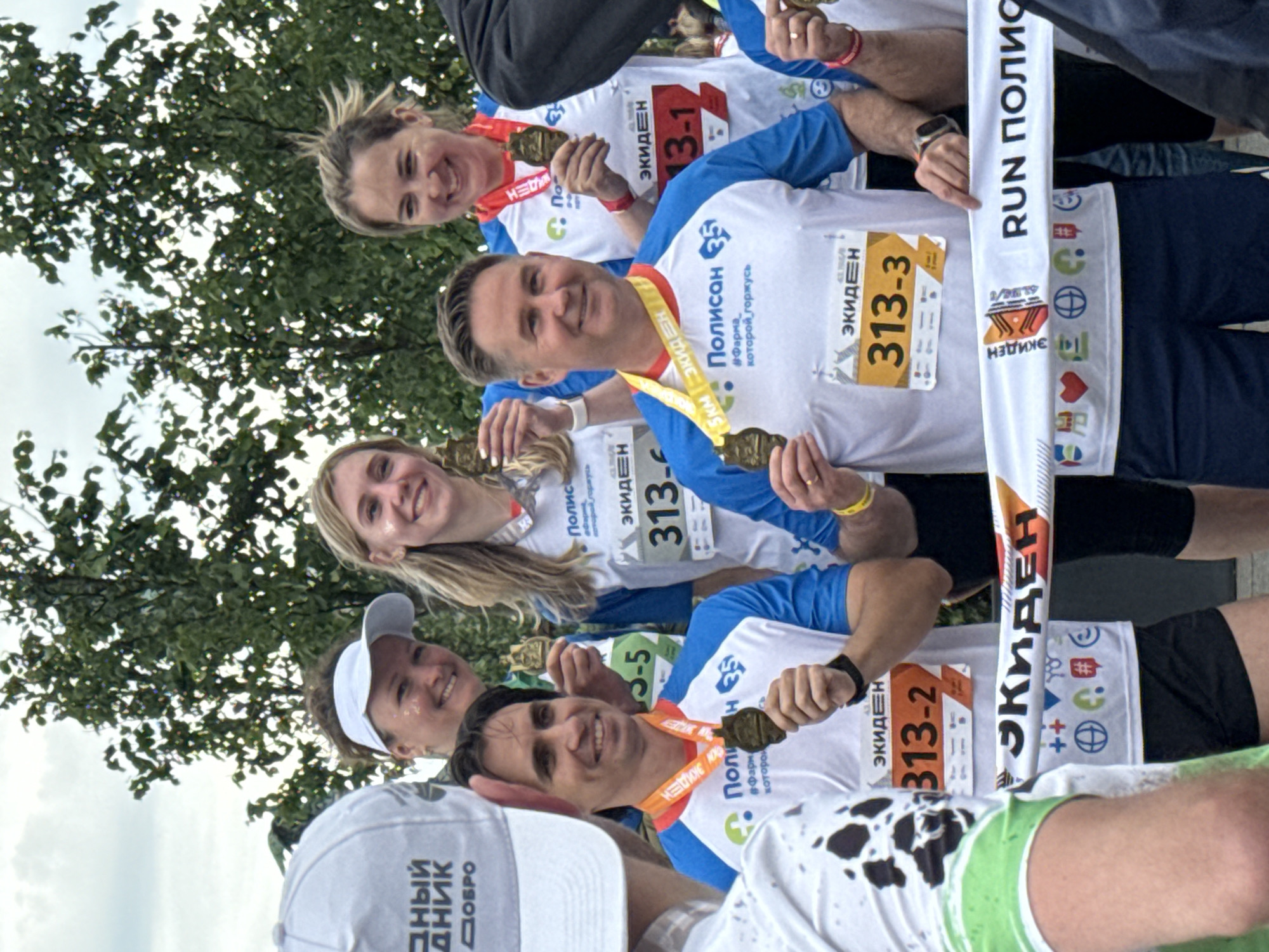 The image depicts a group of smiling individuals, appearing to be runners, posing for a photograph outdoors. They are all wearing matching white t-shirts with blue sleeves and various logos, including "Полисан" and "ЭКИДЕН." Several individuals are wearing medals around their necks, indicating they have participated in a race. One man in the foreground is holding a medal in his hand. Another man is wearing a bib with the number "313-2." Behind them, a white banner with colorful logos and the text "RUN ПОЛИС" is stretched across. The background shows trees and a cloudy sky, suggesting an outdoor event. The event is taking place in Saint Petersburg, Russia, as indicated by the text on the banner and the event's likely association with the "Polisan" company, a pharmaceutical distributor based in Saint Petersburg. The overall atmosphere is celebratory and positive, with participants appearing proud of their accomplishments.