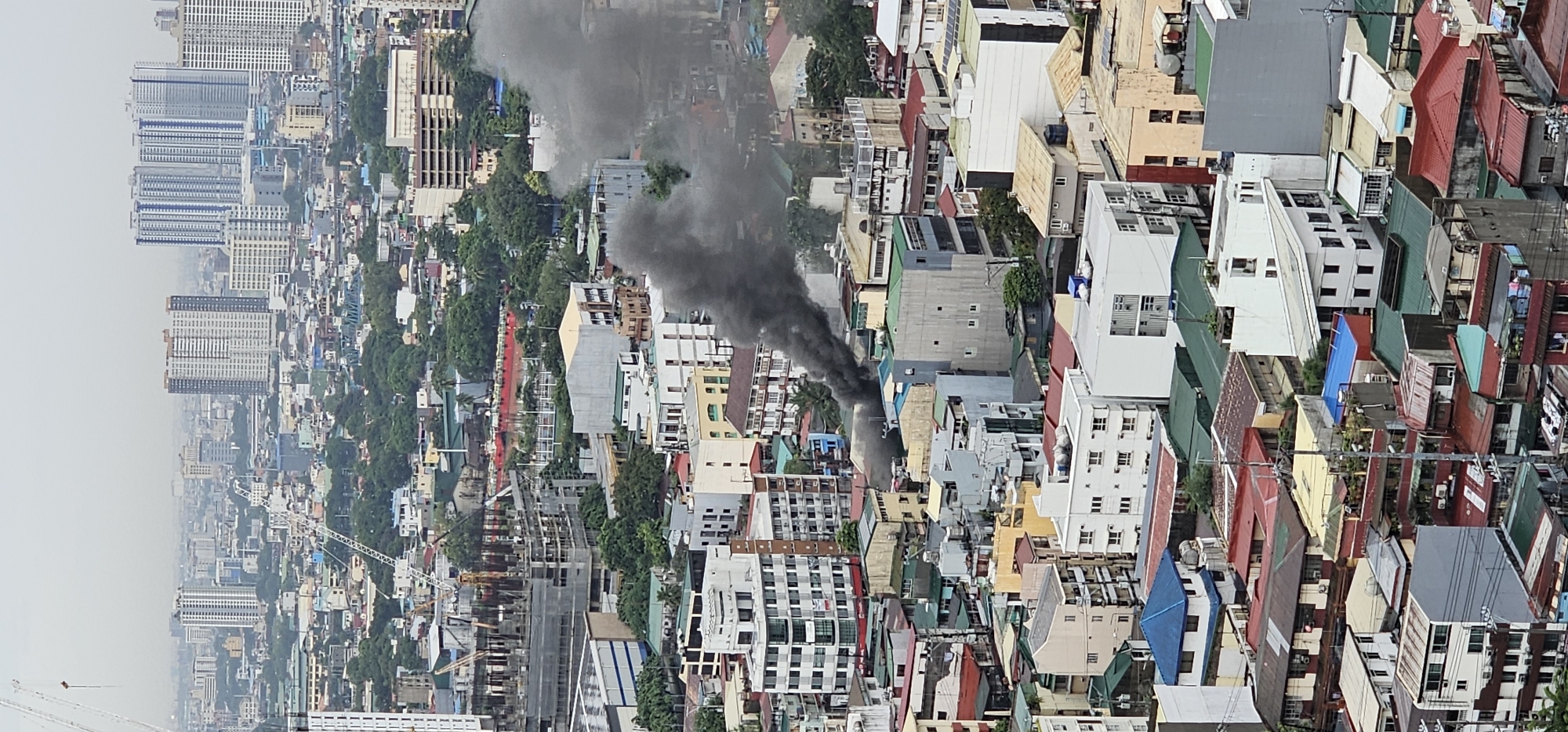 This is a vertical, aerial photograph of an urban landscape, likely taken from a high vantage point, possibly a tall building or drone. The scene depicts a dense residential and commercial area within the city of Makati, Philippines, characterized by a mix of low-rise and mid-rise buildings, interspersed with greenery. In the mid-ground of the image, a significant plume of thick, dark smoke is rising from a cluster of buildings, indicating a fire or other event causing substantial smoke emission. The smoke obscures the buildings from which it originates and drifts upwards into the hazy sky, suggesting it is an active and ongoing incident. The surrounding buildings are varied in their architectural styles and roof colors, predominantly featuring red, green, and white facades. Cranes for ongoing construction are visible in the background, alongside taller, more modern high-rise buildings that define the Makati skyline. The overall atmosphere is overcast, with a muted color palette.