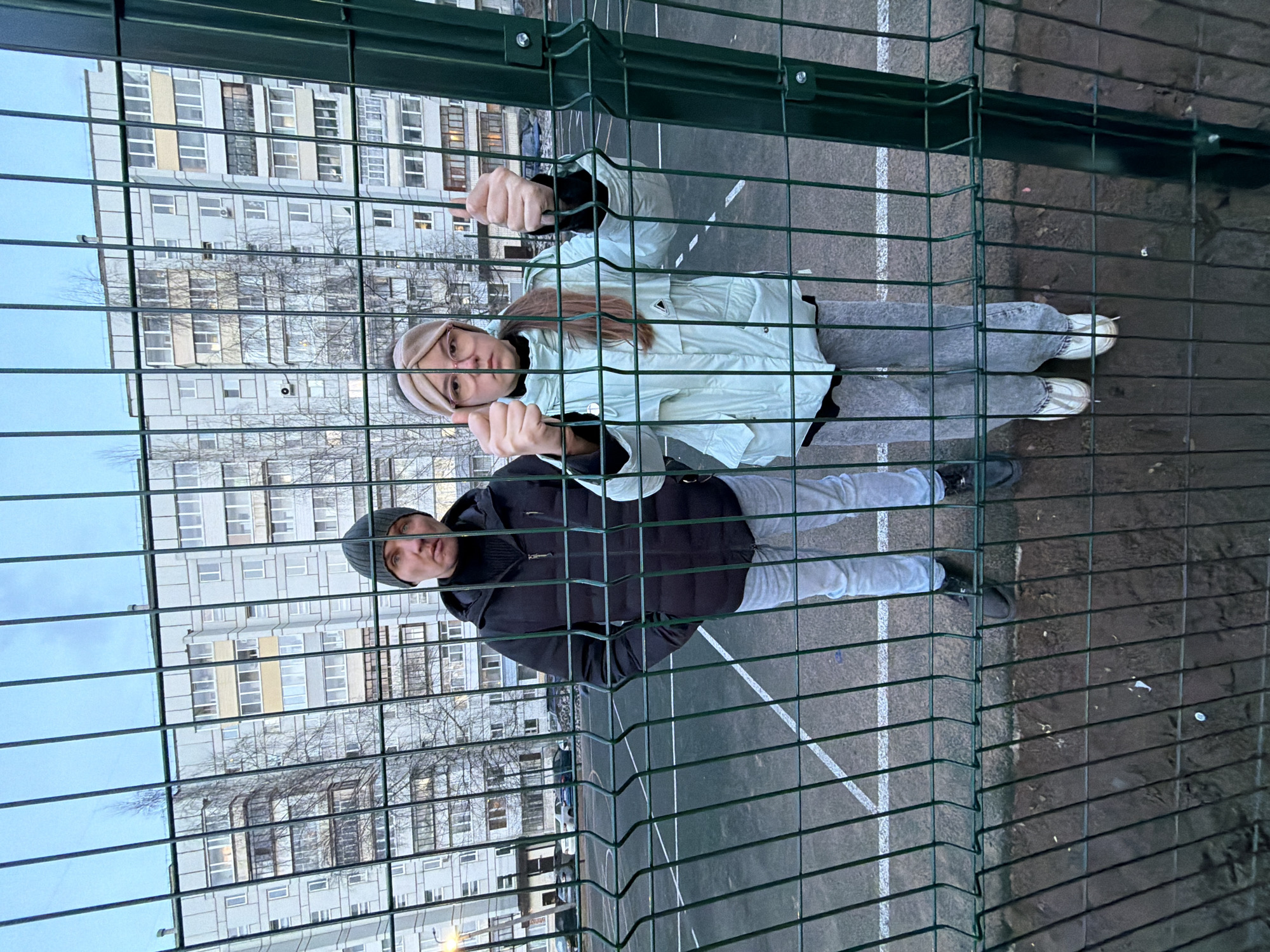 This image captures an outdoor scene featuring two individuals posing behind a green metal mesh fence. The setting appears to be a sports court or playground, indicated by the paved ground with white markings. In the background, multi-story residential buildings with numerous windows are visible under an overcast sky, suggesting an urban or suburban environment. Bare trees between the buildings and the fence indicate cool weather, likely late autumn or winter.
The person on the left is a female with long, light brown hair, wearing glasses and a light-colored headband. She is dressed in a light mint green or pale blue puffer jacket, a black top, light grey jeans, and white sneakers. She holds onto the fence with both hands, giving a thumbs-up gesture. Her expression is calm, with a slight smile.
To her right stands a male, wearing a dark grey or black beanie, a black puffer jacket over a black top, and light grey pants with dark shoes. He is also holding onto the fence with his right hand. His expression is neutral.
The individuals appear to be aware of the camera, posing for the photograph. The time of day is daytime, but the soft, diffused lighting suggests an overcast sky or late afternoon. No visible text is present in the image. The location is Chaikovsky, Russia.