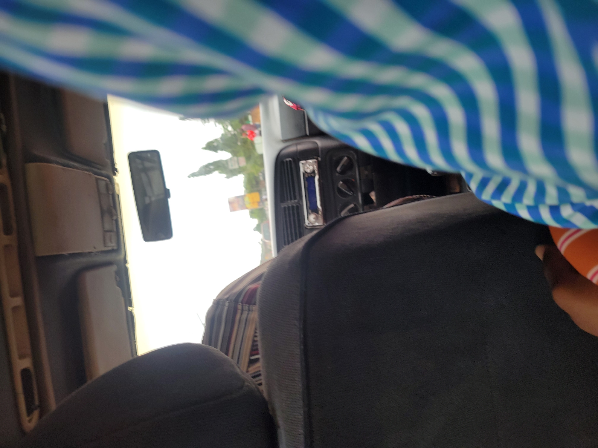 The image shows an interior view of a vehicle, possibly a car or truck, in Unknown, Unknown. The interior is tan and black. A rearview mirror is visible. The front dashboard has a radio unit with buttons. Outside the window, trees and signage are visible. The image also captures part of a person's shirt, which has a blue and white checkered pattern. A hand with an orange object is also visible.