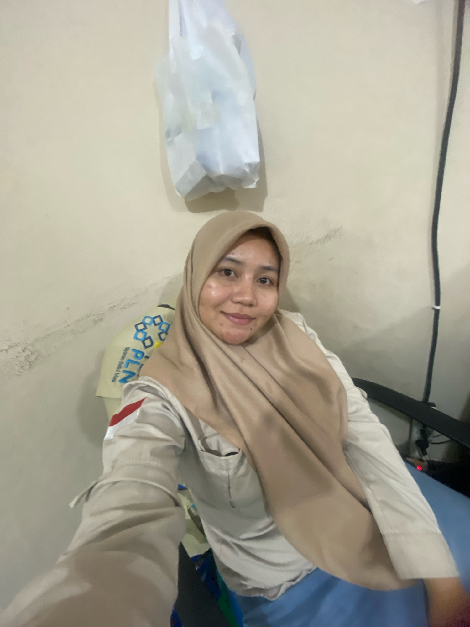 A woman wearing a tan hijab and a tan uniform shirt with a red and white flag patch on the sleeve is sitting in a chair. The uniform shirt has "PLN" written on it in blue and white letters. The background is a plain, light-colored wall. A white plastic bag is hanging from the wall behind the woman. A black cable is also visible on the wall. The overall setting appears to be indoors, possibly an office or a residential room, located in Banjarbaru, Indonesia.
