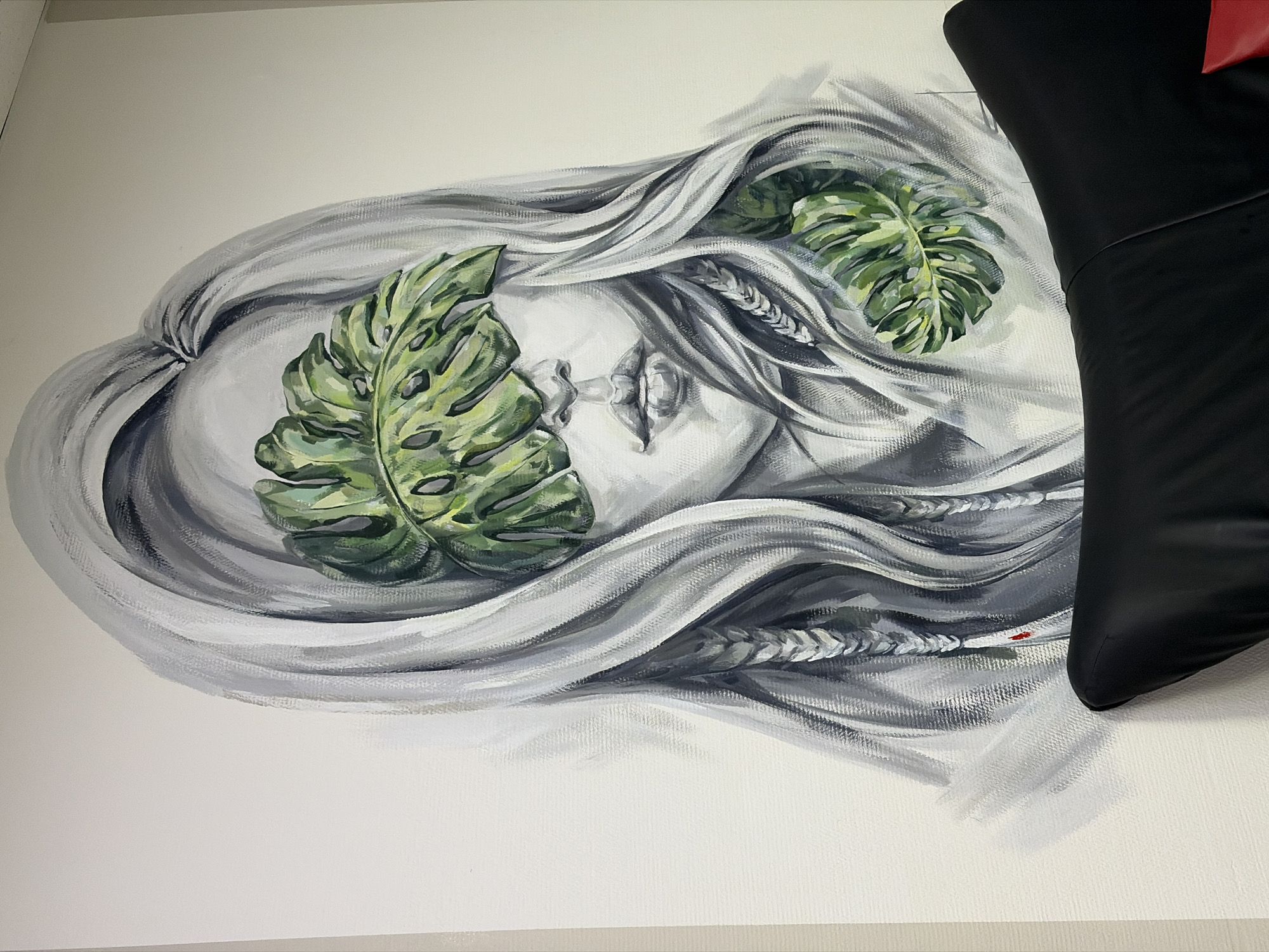 The image shows a painting on a white wall. The painting depicts a woman's face in profile, with flowing grey hair interwoven with two large monstera leaves. The woman's eyes are closed, and her lips are slightly parted. Her hair also contains sections that resemble tightly woven braids. The painting style is realistic, with careful attention to shading and texture. A dark, partially obscured object, possibly part of a chair, is positioned to the right of the painting, partially obscuring the right side of the artwork. The context lacks sufficient details to identify the precise location in Chaikovsky, Russia.