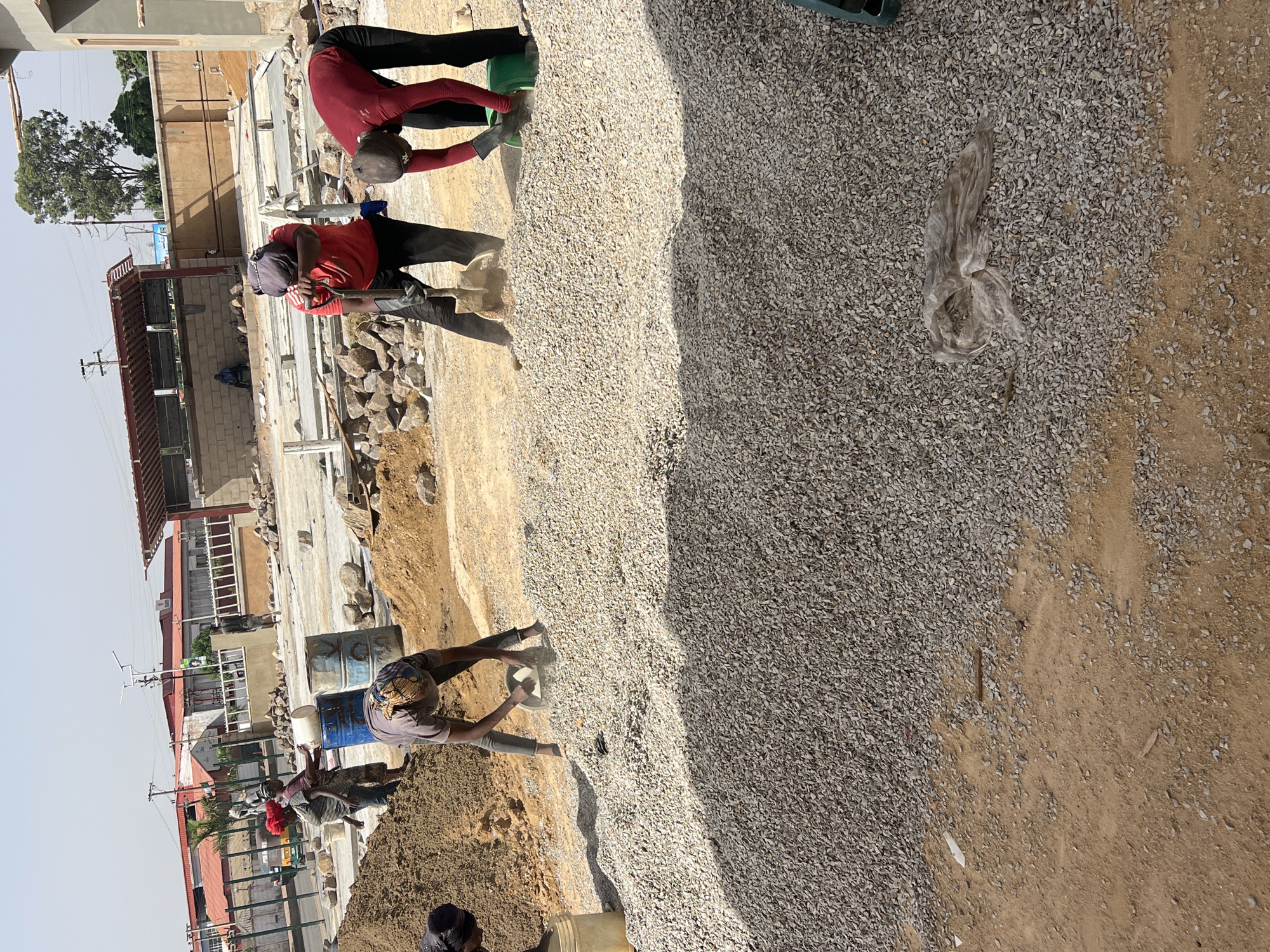 The image captures a construction site in Jos, Nigeria, featuring several individuals engaged in manual labor. Two workers in red shirts and dark trousers are using shovels to move gravel. Another person is pouring water from a white bucket into a blue barrel. Buildings are visible in the background, along with a partially constructed metal structure. The foreground is dominated by a large pile of gravel, next to a dirt area.