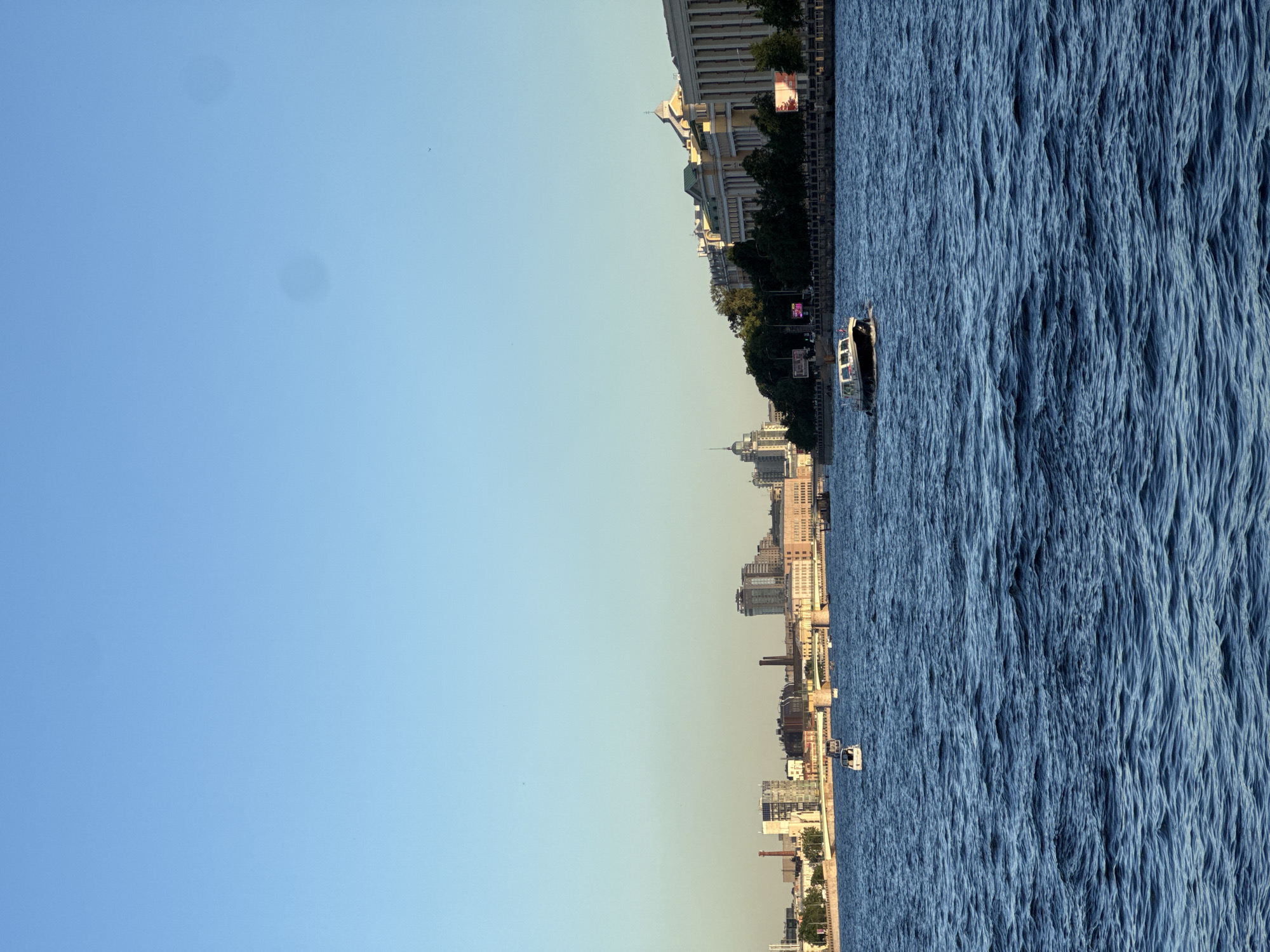 The image shows a wide river in a city with a clear sky and buildings along the embankment. The river is blue and has noticeable waves. There are several boats on the water, including a small white motorboat in the foreground, a larger white boat with a dark roof moving towards the right, and another smaller boat further in the distance. The embankment features a row of buildings of varying architectural styles, some appearing older and classical, others more modern. Trees line the embankment in front of some of the buildings. A bridge spans the river in the background. The sun appears to be low in the sky, casting a warm light on the scene.