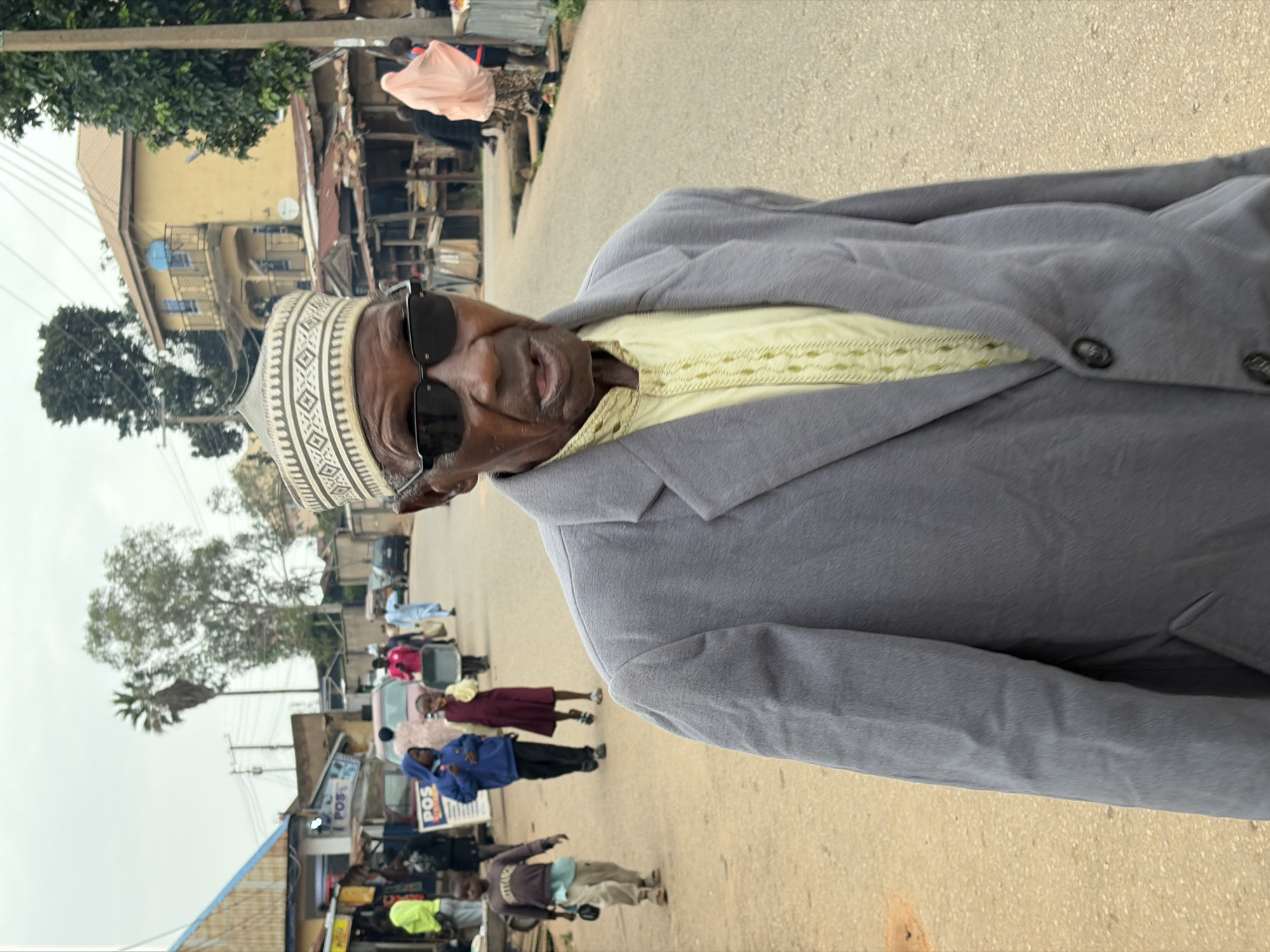 An elderly man wearing a gray suit jacket, a light yellow patterned shirt, and a white and black patterned cap, stands in the foreground of a street in Jos, Nigeria. He is also wearing dark sunglasses. Behind him, several people, including children, walk along the dusty road. Shops line the sides of the street, with some displaying signs. A yellow building with balconies is visible in the background, along with trees and a cloudy sky. The man's mouth is slightly open, suggesting he may be speaking. The scene appears to be a typical day in an urban area.