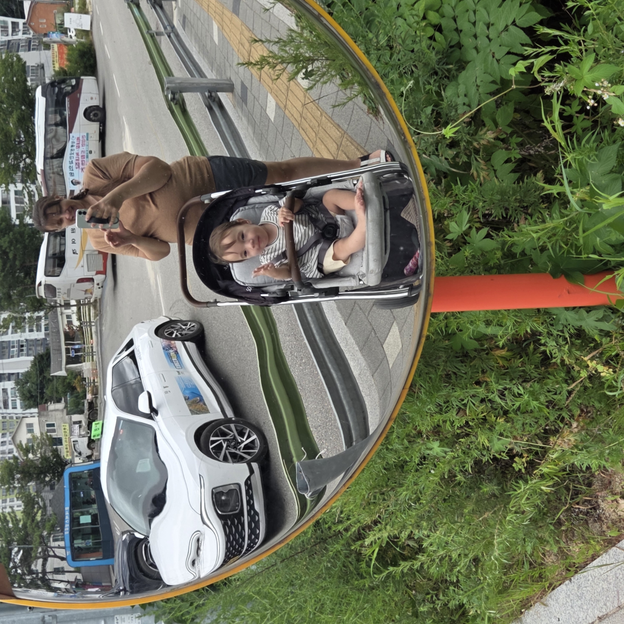 The image shows a reflection in a convex mirror, likely a traffic mirror, situated in Dunnae-myeon, South Korea. In the reflection, a woman is holding a smartphone and taking a picture of herself and a toddler in a stroller. The toddler, wearing a striped shirt, is seated in a grey stroller and appears to be gesturing. The woman is wearing a brown shirt and denim shorts. Behind them, a white taxi with advertising on its side is visible, along with other vehicles, including a dark SUV and a bus. The background consists of buildings and trees, suggesting an urban or suburban environment. The mirror is mounted on a red pole and surrounded by green foliage. The ground in the reflection shows a sidewalk with tactile paving.