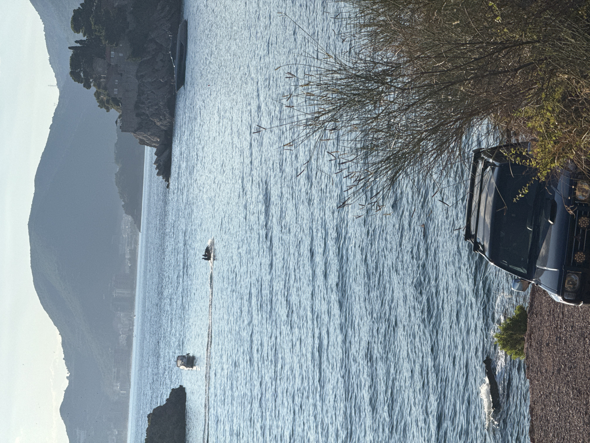 The image depicts a scenic view of the Adriatic Sea near Sveti Stefan, Montenegro. The water is a vibrant blue, with ripples suggesting a light breeze. In the foreground, the top of a dark-colored vehicle is visible, parked on a gravelly embankment. The vehicle appears to be an off-road or utility type, with visible headlights. To the left of the water, a rocky cliff rises, topped with lush green trees and a building complex. Further in the distance, a hazy, mountainous landscape stretches across the horizon. Two small boats are present on the water. One is closer to the cliff, and another is further out, leaving a distinct wake. The time of day appears to be late afternoon or early evening, indicated by the soft, diffused sunlight. The overall mood is tranquil and picturesque, capturing the natural beauty of the Montenegrin coast.