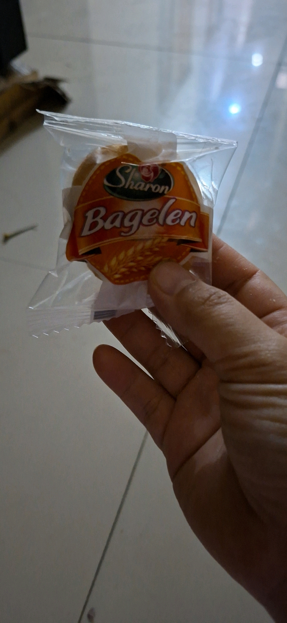 A person's hand is holding a clear plastic package containing a single, round baked good. The package has a colorful label with orange and yellow hues. Prominently displayed on the label is the brand name "Sharon" in dark script, with a stylized red rose above it. Below "Sharon," the word "Bagelen" is written in large, white, sans-serif font, with wheat stalks depicted at the bottom of the label. The background appears to be an indoor floor, possibly tiled, with some lighting reflections visible. The image is taken from a slightly high angle, looking down at the hand and the packaged item. There are no specific identifying landmarks visible to pinpoint a location within Surabaya, Indonesia.