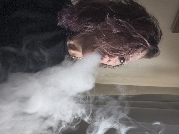 This image depicts an indoor scene in what is reported to be the city of Westminster, United States of America. A single adult individual is shown in a close-up, chest-up shot, viewed from a slightly right-of-center perspective. The subject has messy, medium-length hair with purple and pinkish hues, partially obscuring the right side of their face. The visible left eye features dark eyeliner and is directed forward. A substantial plume of dense, white vapor or smoke is being exhaled from the individual's mouth, extending diagonally across the lower right quadrant of the frame. A thin silver-colored chain necklace is visible around the neck, and a dark, angular tattoo is present on the left side of the neck, beneath the jawline. The individual is wearing a plain, dark-colored crew-neck garment. The background consists of a light-colored wall or curtain on the left, with a faint, dark rectangular object (possibly a framed item) on the upper left. To the right, a darker, semi-transparent curtain or window covering is visible.