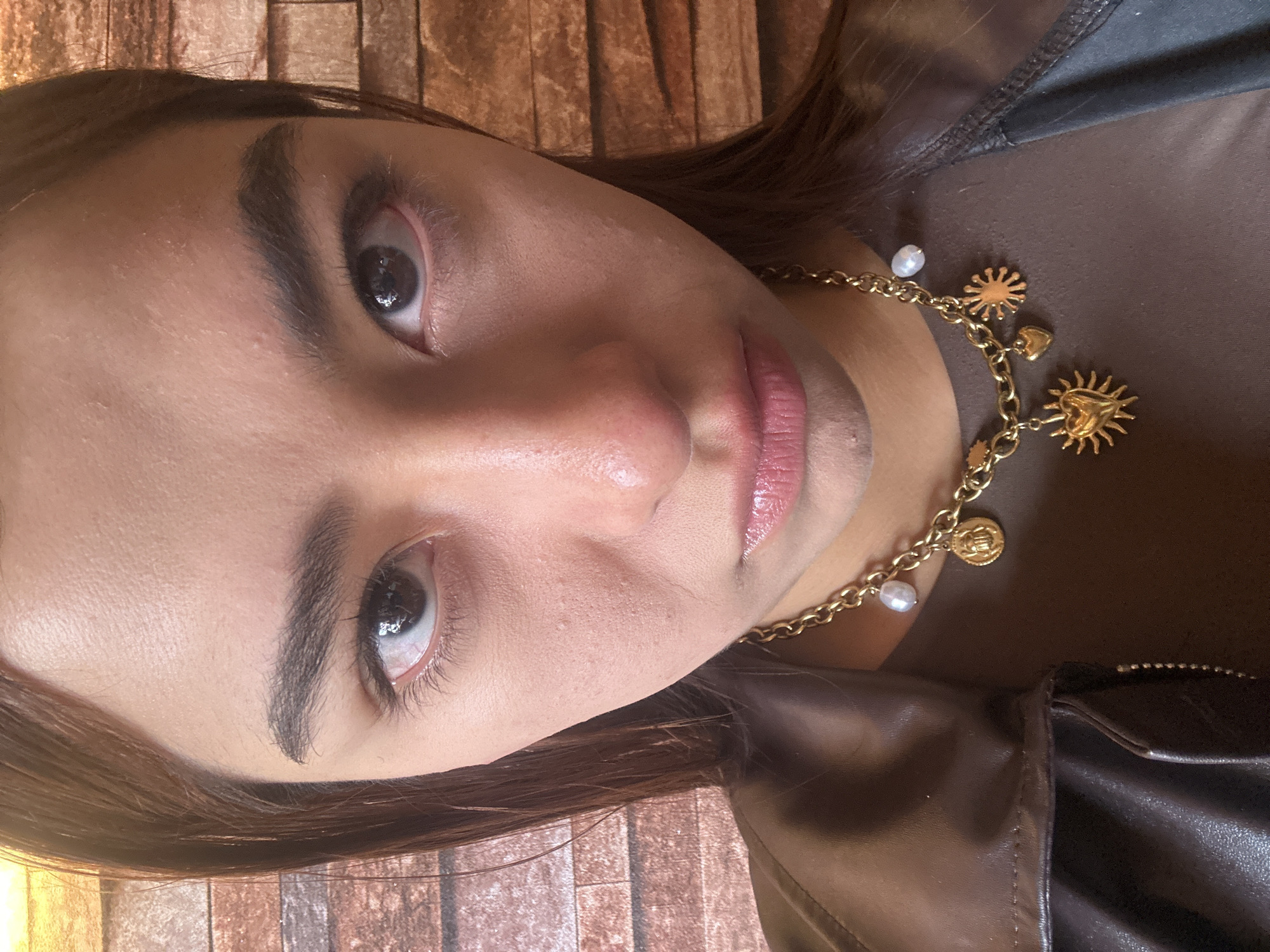 A close-up portrait of a woman is oriented vertically. She has dark eyebrows, brown hair, and light skin. She wears light pink lipstick. A gold necklace with charms and pearls is visible around her neck, partially obscured by her clothing. She wears a brown top and a dark leather jacket. The background appears to be a brick wall. The location is not identifiable from the image.